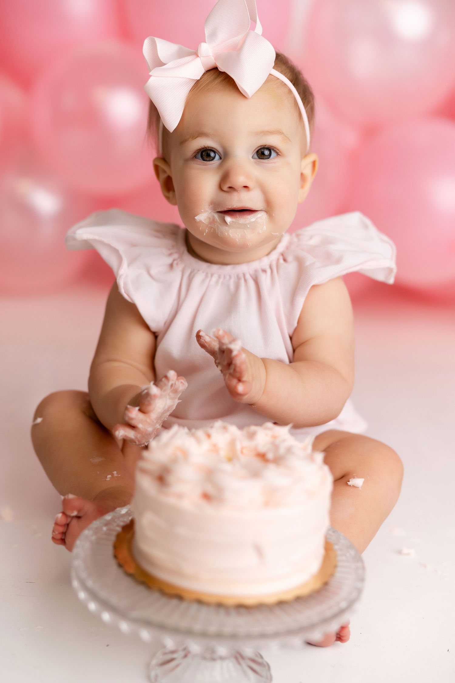Baby in pink dress and bow sits with smiling expression next to white frosted cake during cake smash photo session.