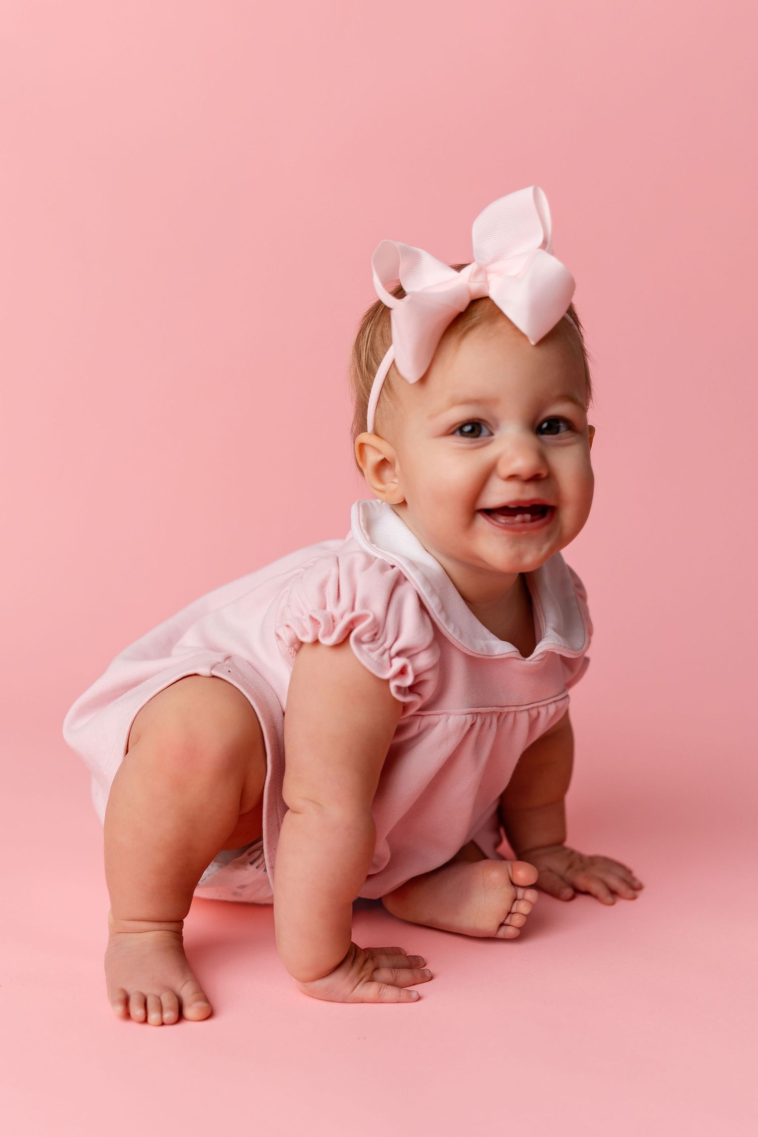 Baby in pink dress and bow crawls on pink background during portrait session.