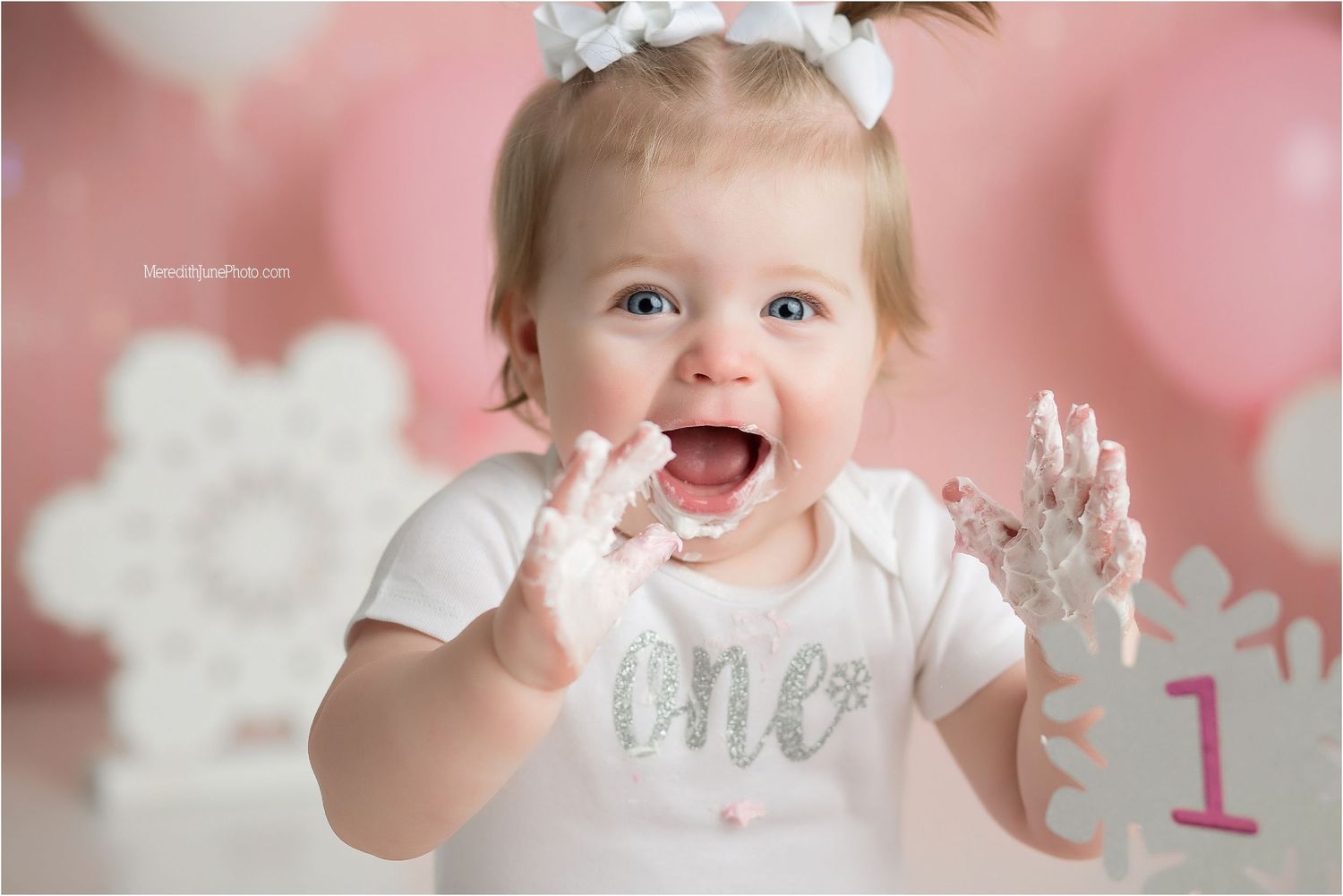Baby in white dress celebrates first birthday with pink balloons and silver decorations in background.