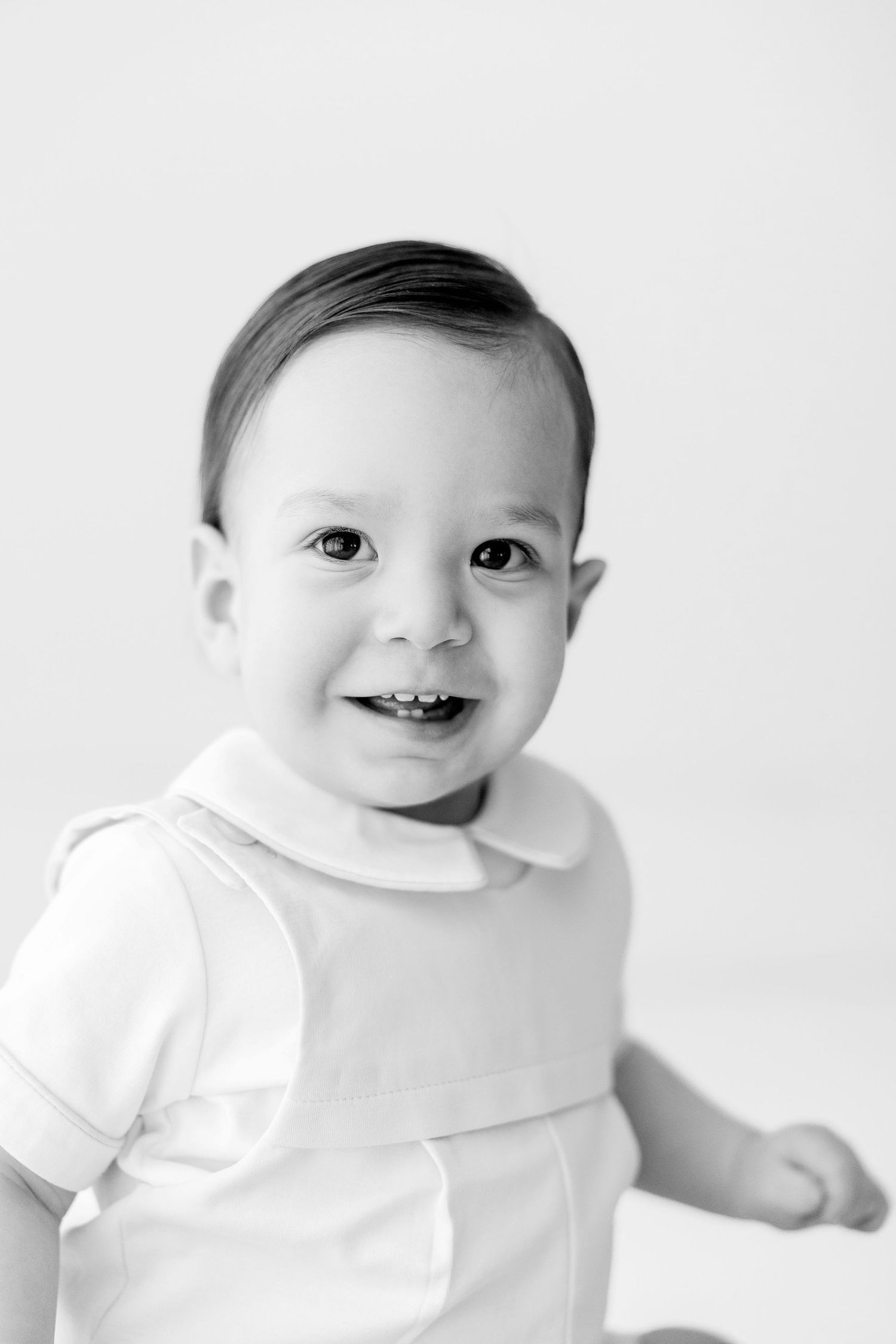Black and white portrait of a smiling baby wearing a white outfit against a light background.