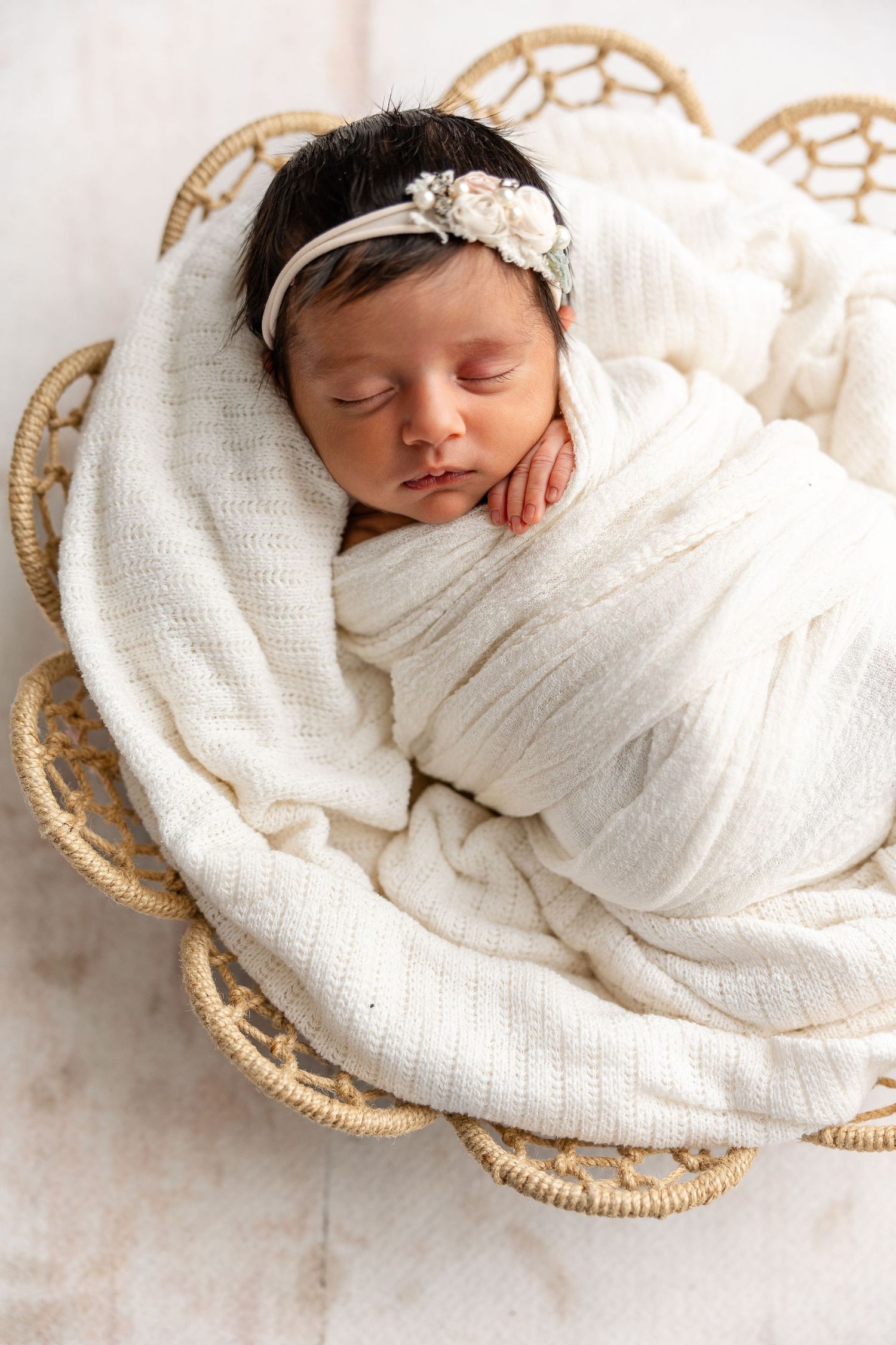 Sleeping newborn baby wrapped in white swaddle with floral headband rests in woven basket.