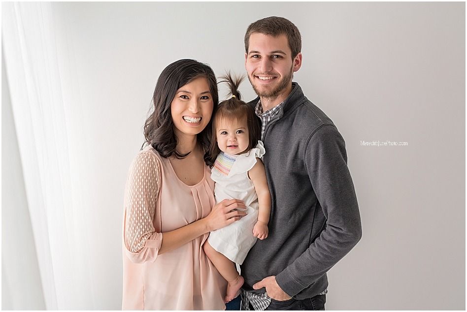 A family poses together against a neutral wall, with a baby wearing light blue clothing in the center.