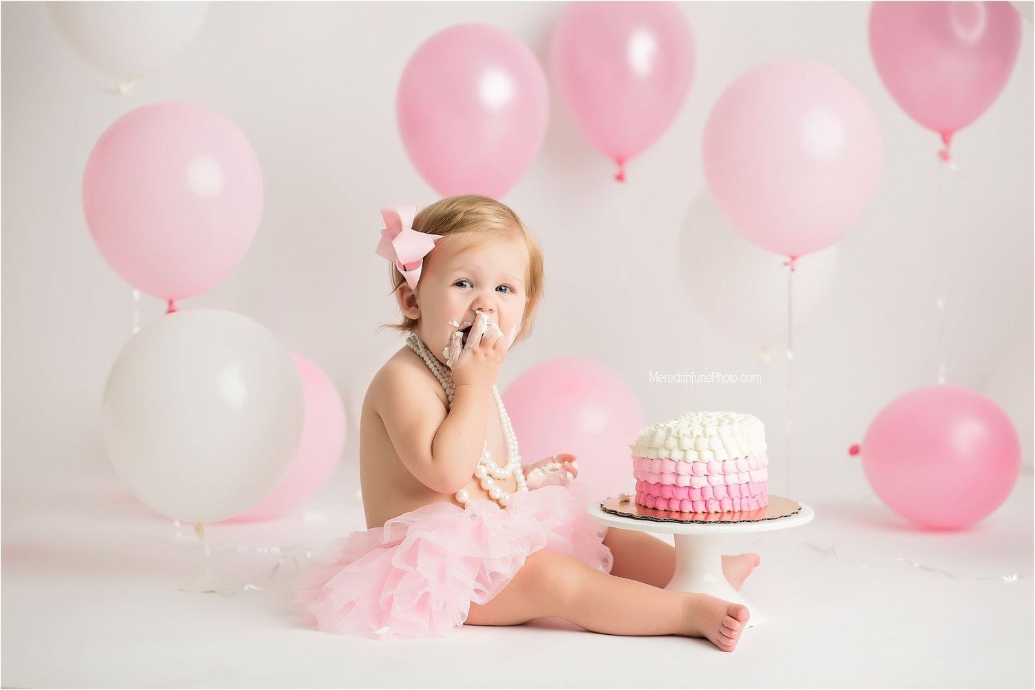Baby in pink dress celebrates first birthday with cake and pink balloons during photo session.