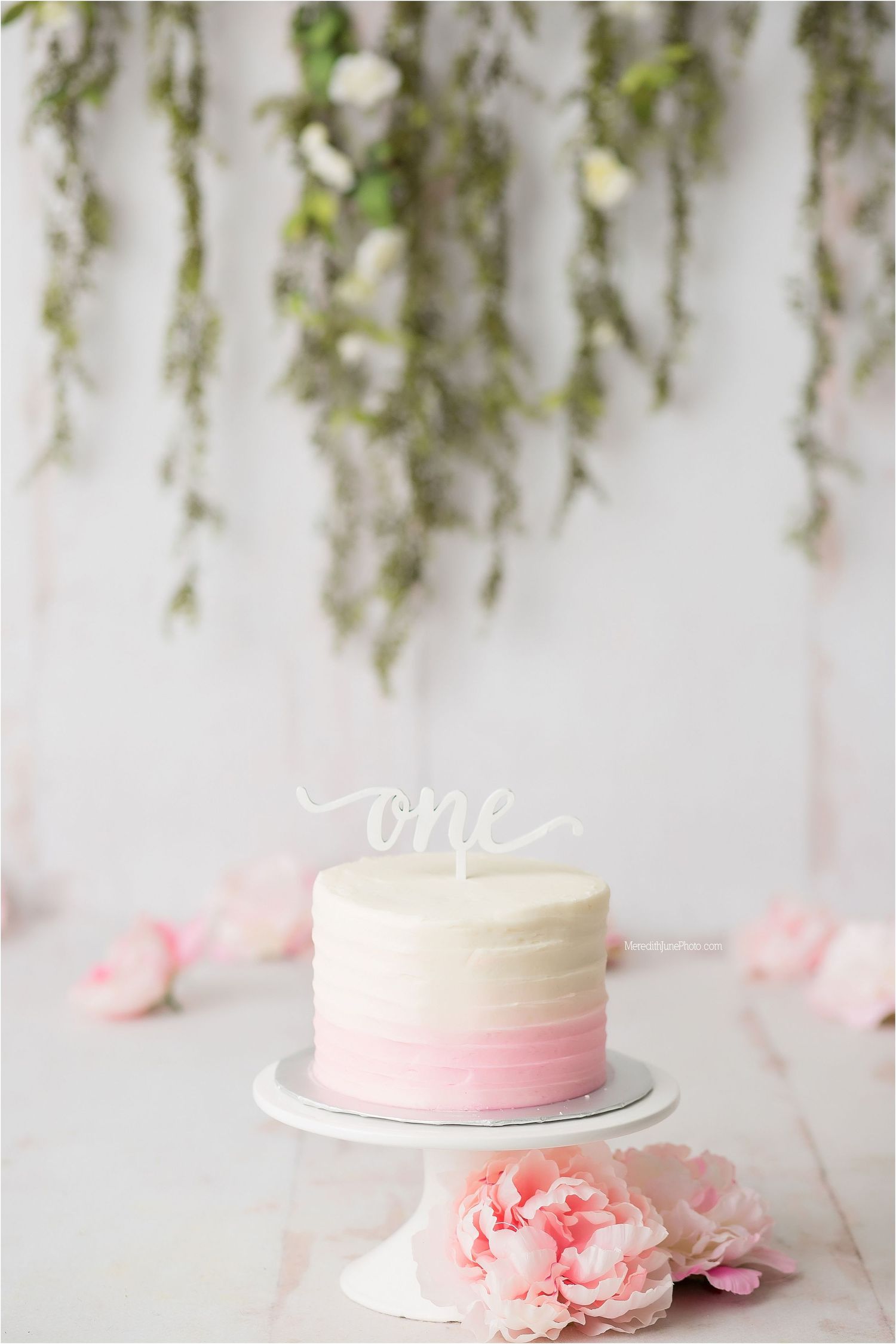 White wedding cake with pink ombre design and delicate floral decorations sits elegantly on white cake stand.