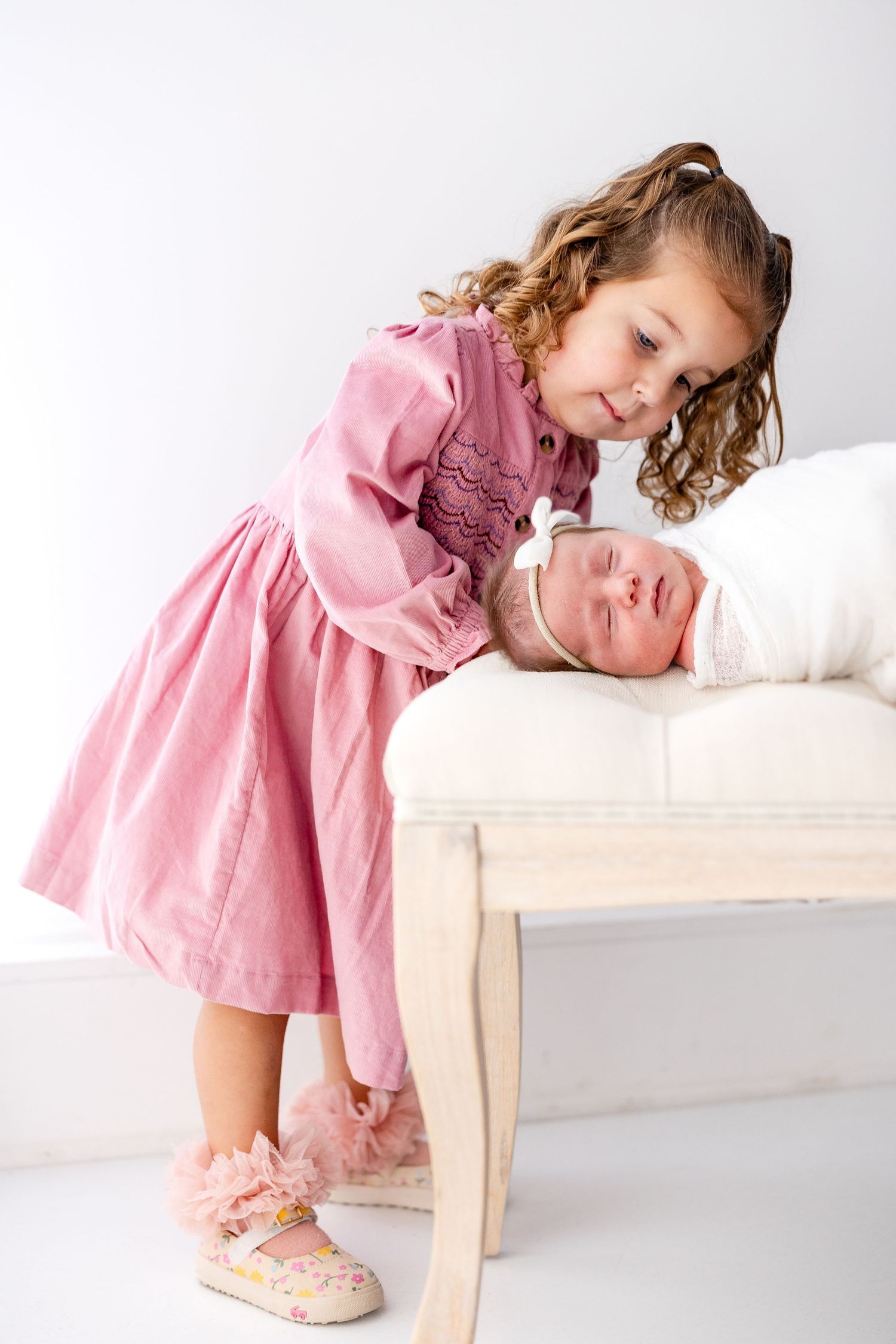 A child in a pink dress leans over white bedding while playing and smiling.