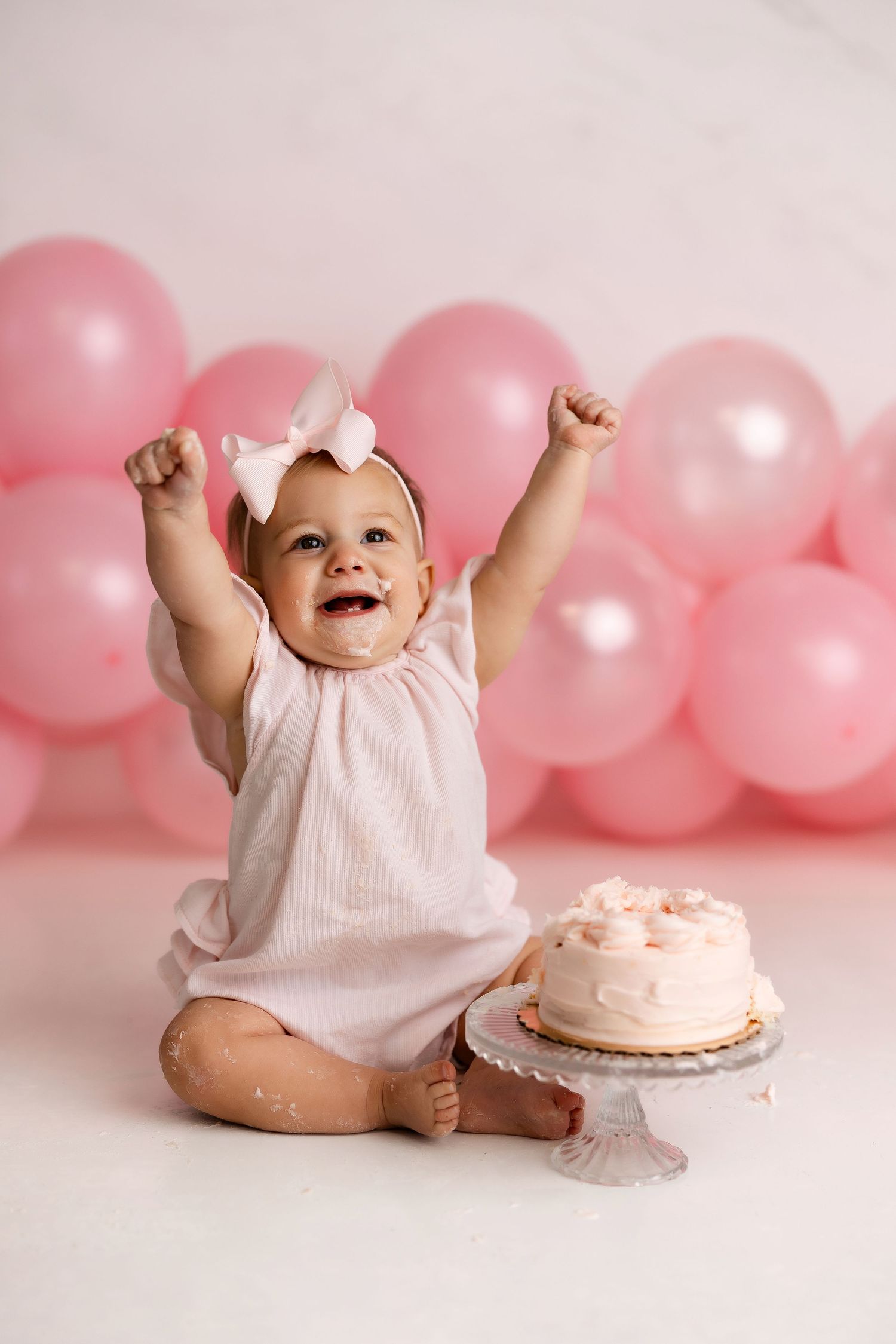 Joyful baby in white dress celebrates first birthday with pink balloons and matching cake on white backdrop.