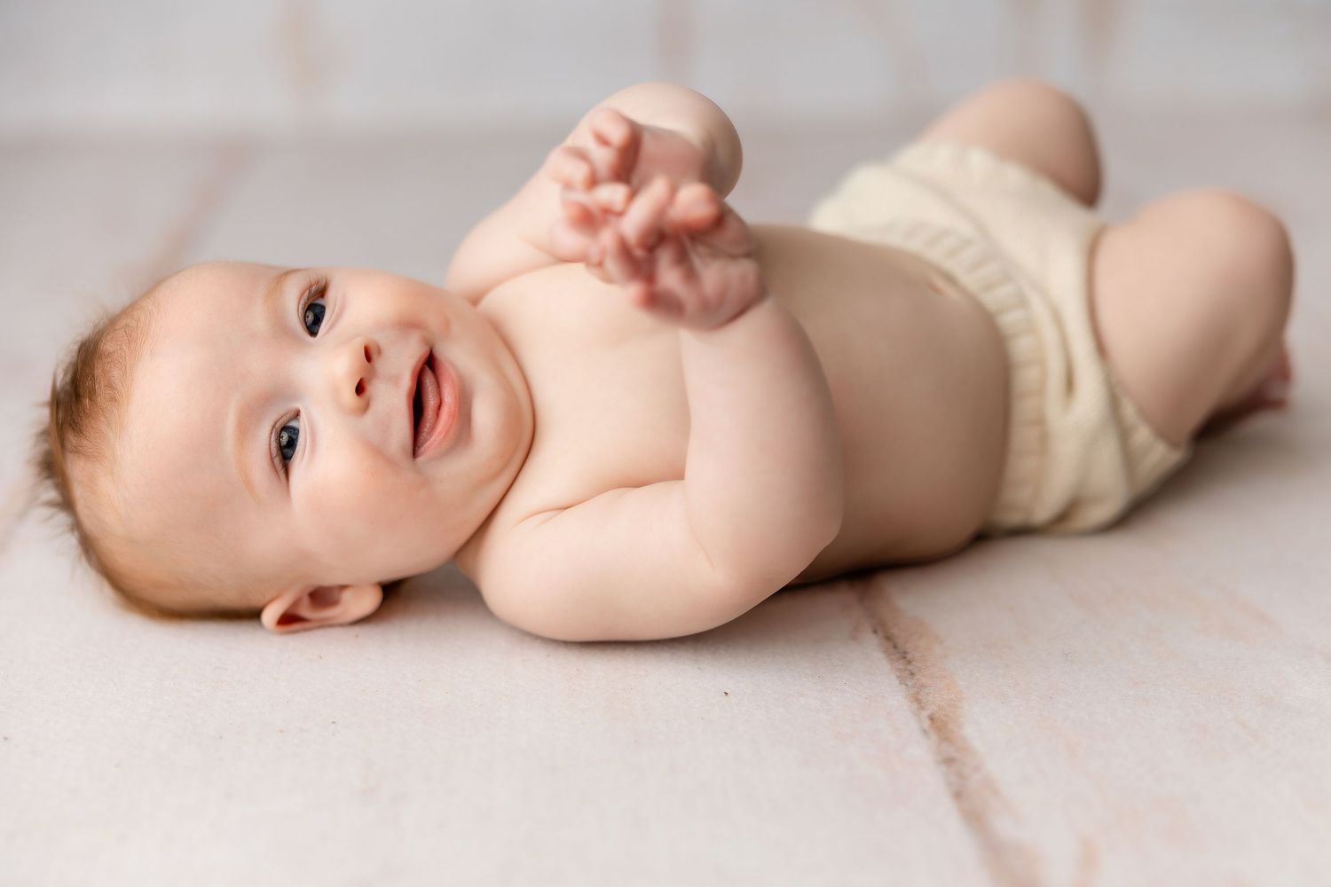 A happy baby in a cream-colored diaper lies on their back on a light colored floor.