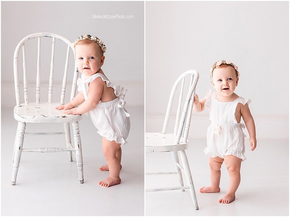 A baby in a white outfit sits and plays on a vintage white wooden chair in a minimalist studio setting.