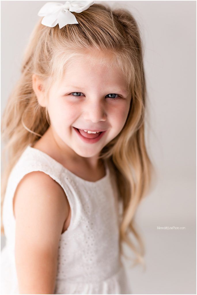 Portrait of a young child wearing a white dress and bow smiling brightly against a light background.