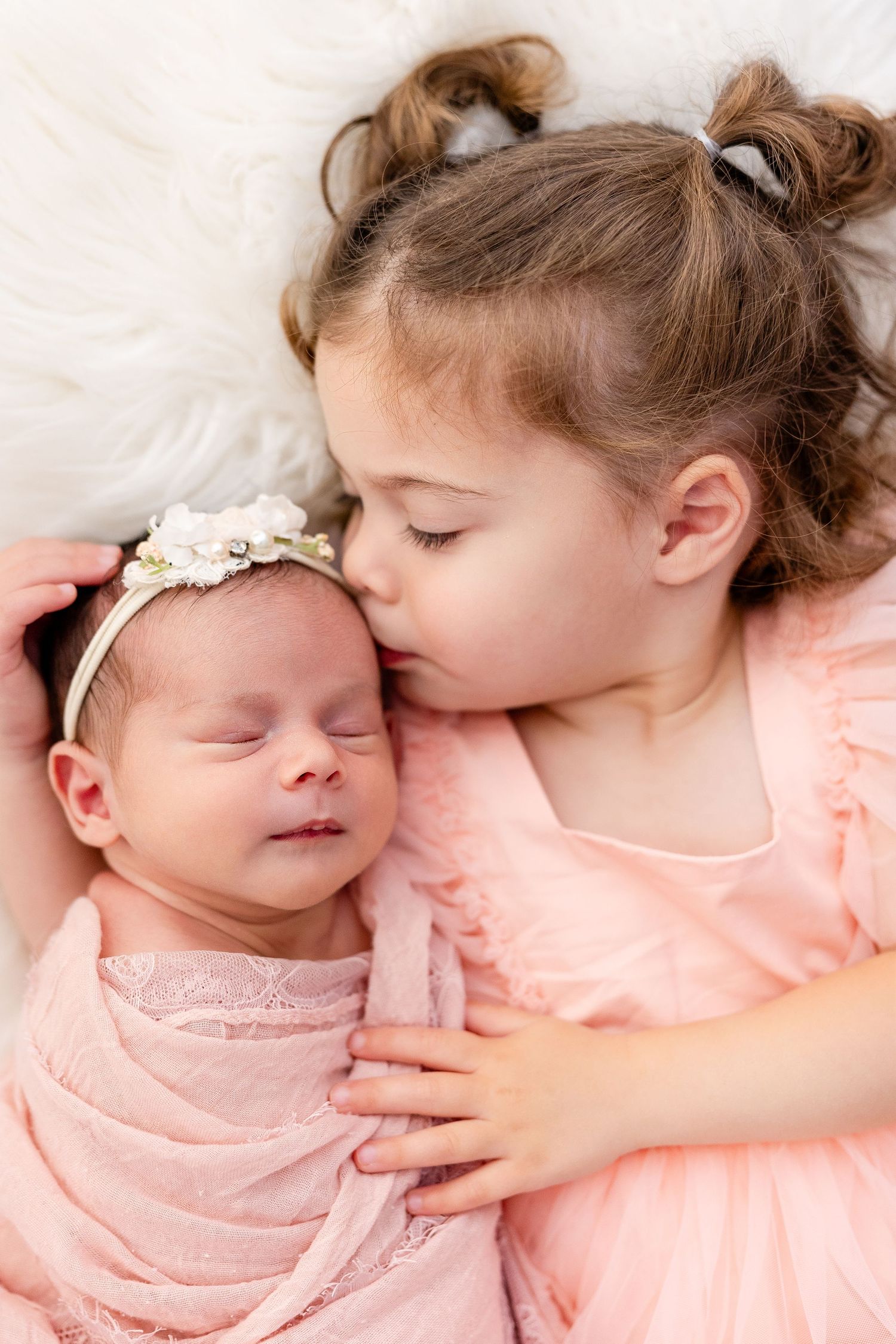 Sweet moment of siblings in pink dresses sharing tender moment with sleeping baby.