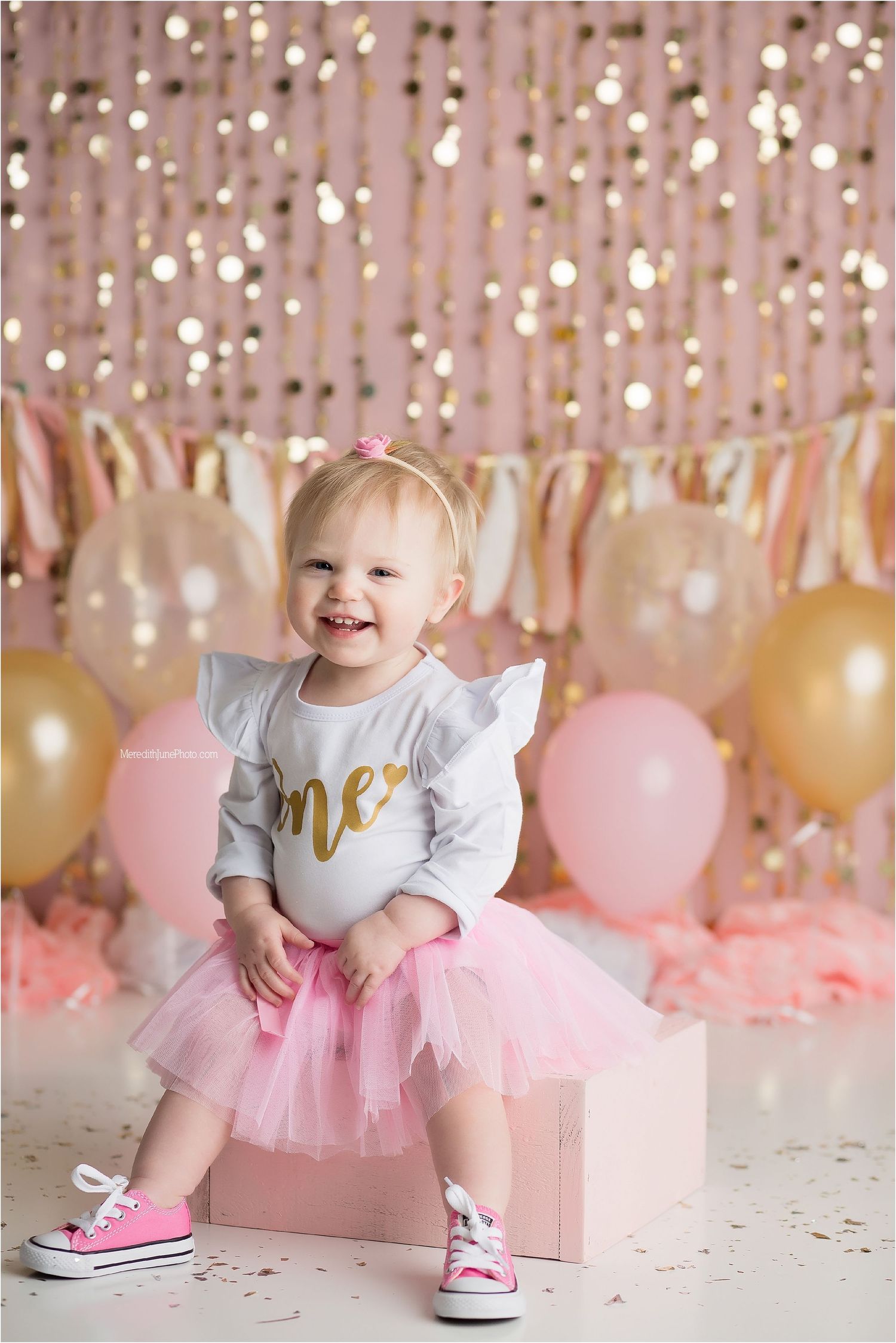 Baby in pink tutu and gray shirt sits on floor with pink and gold balloon decorations for first birthday celebration.