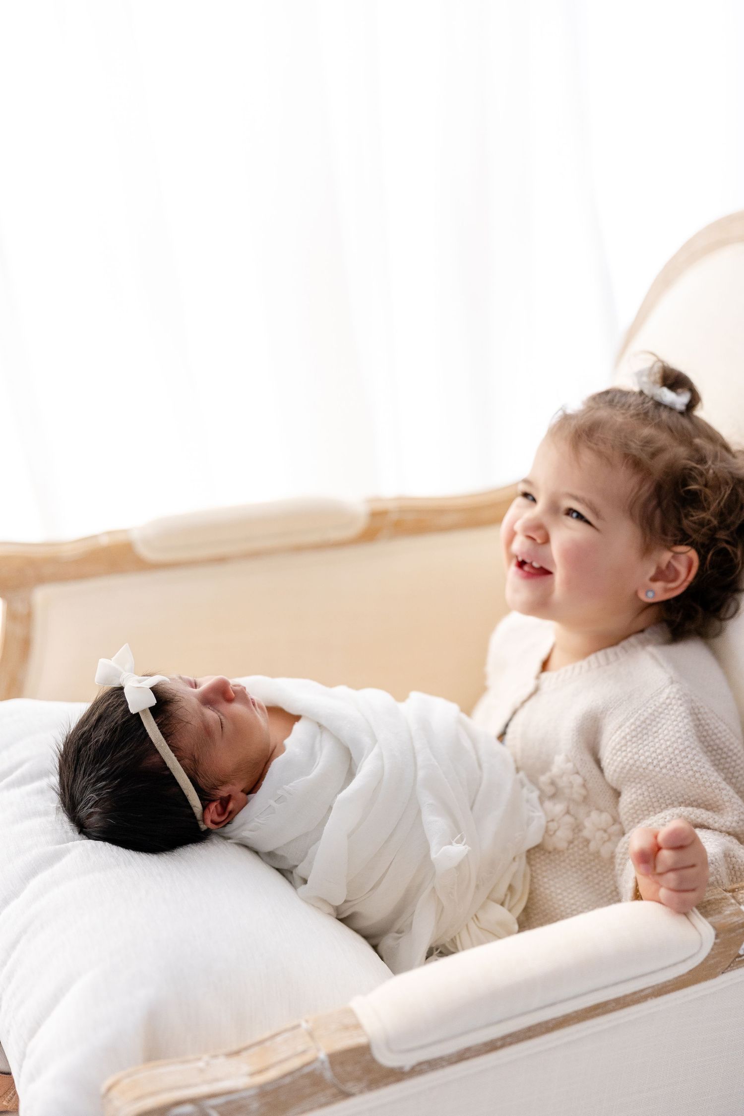 Two children share a sweet moment on a white couch, one sitting up and one lying down.