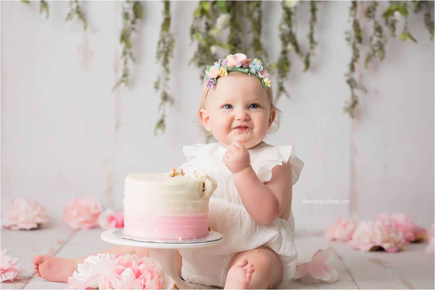 A pink and white cake smash photo session featuring a baby in a tulle dress against a floral backdrop.