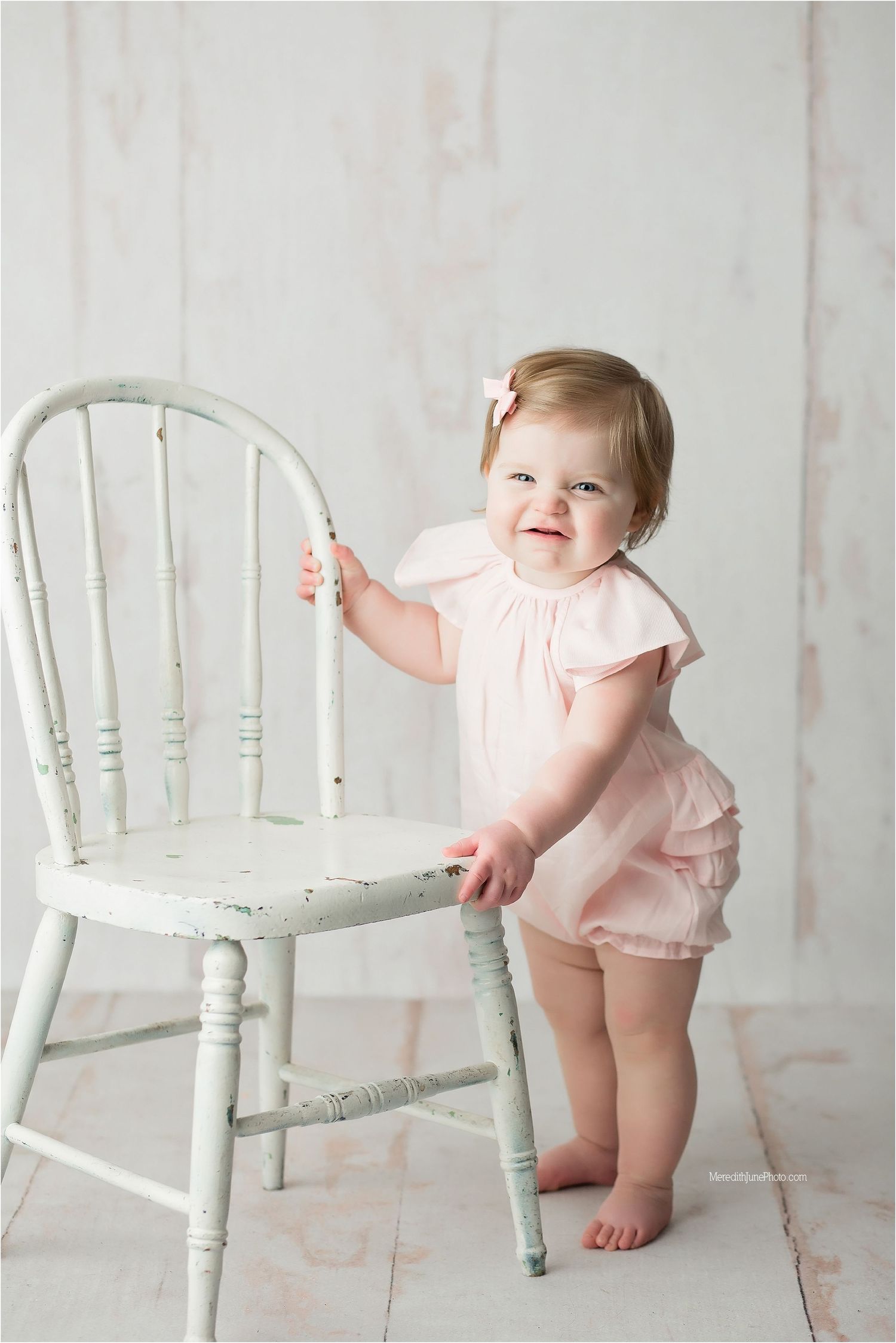Baby in pink romper plays around white vintage chair in studio setting with soft lighting and neutral backdrop.