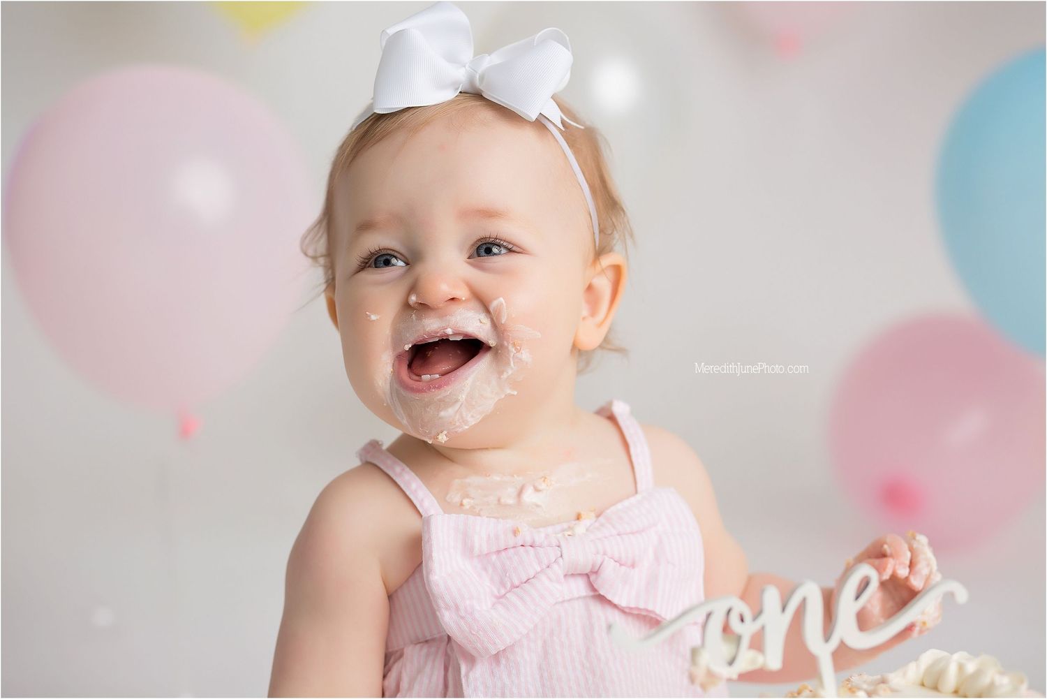 A happy baby wearing pink with a white bow poses against a pastel balloon background.