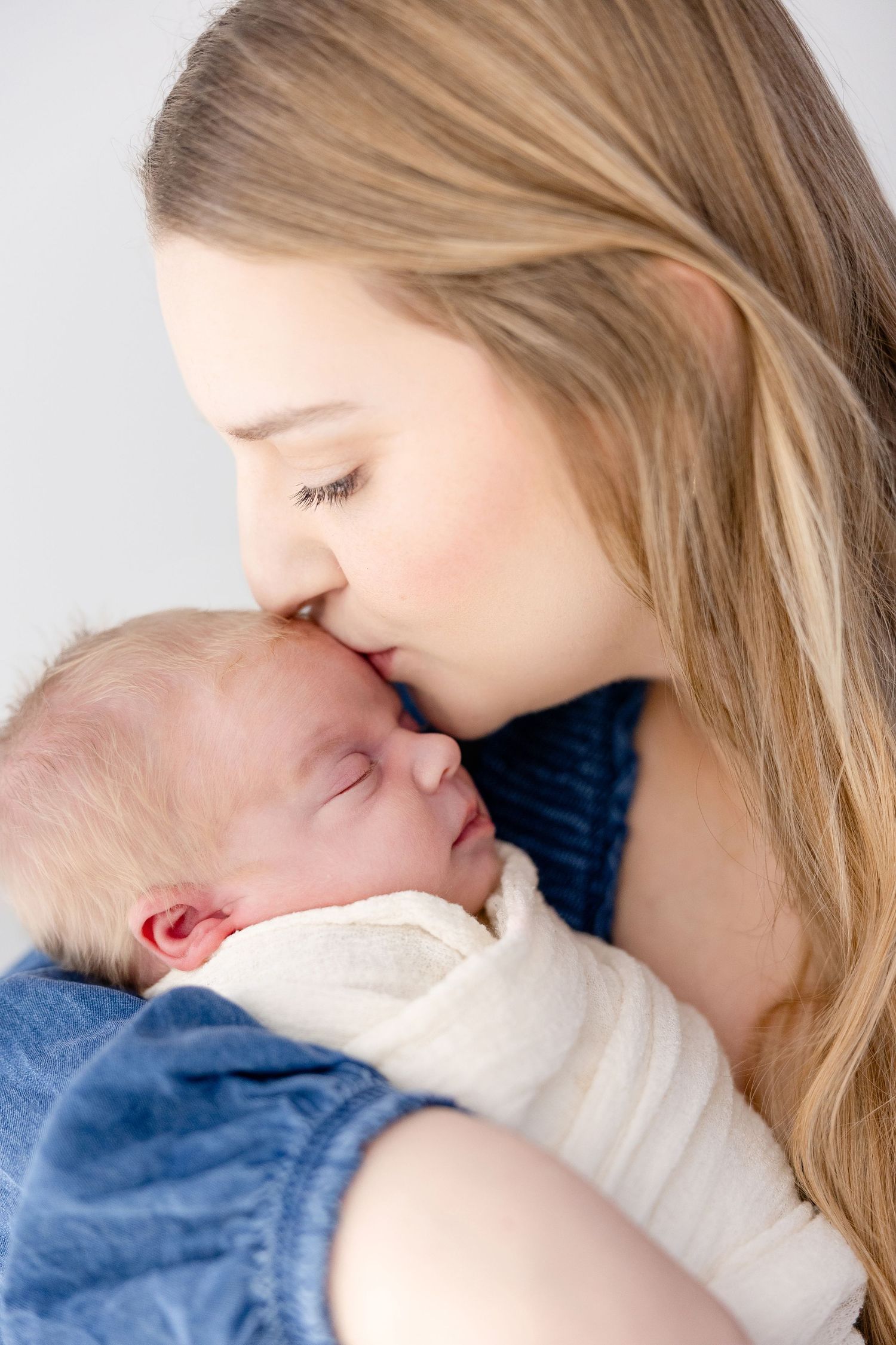 A tender moment captured as a mother gently kisses her sleeping newborn baby wrapped in white and blue blankets.