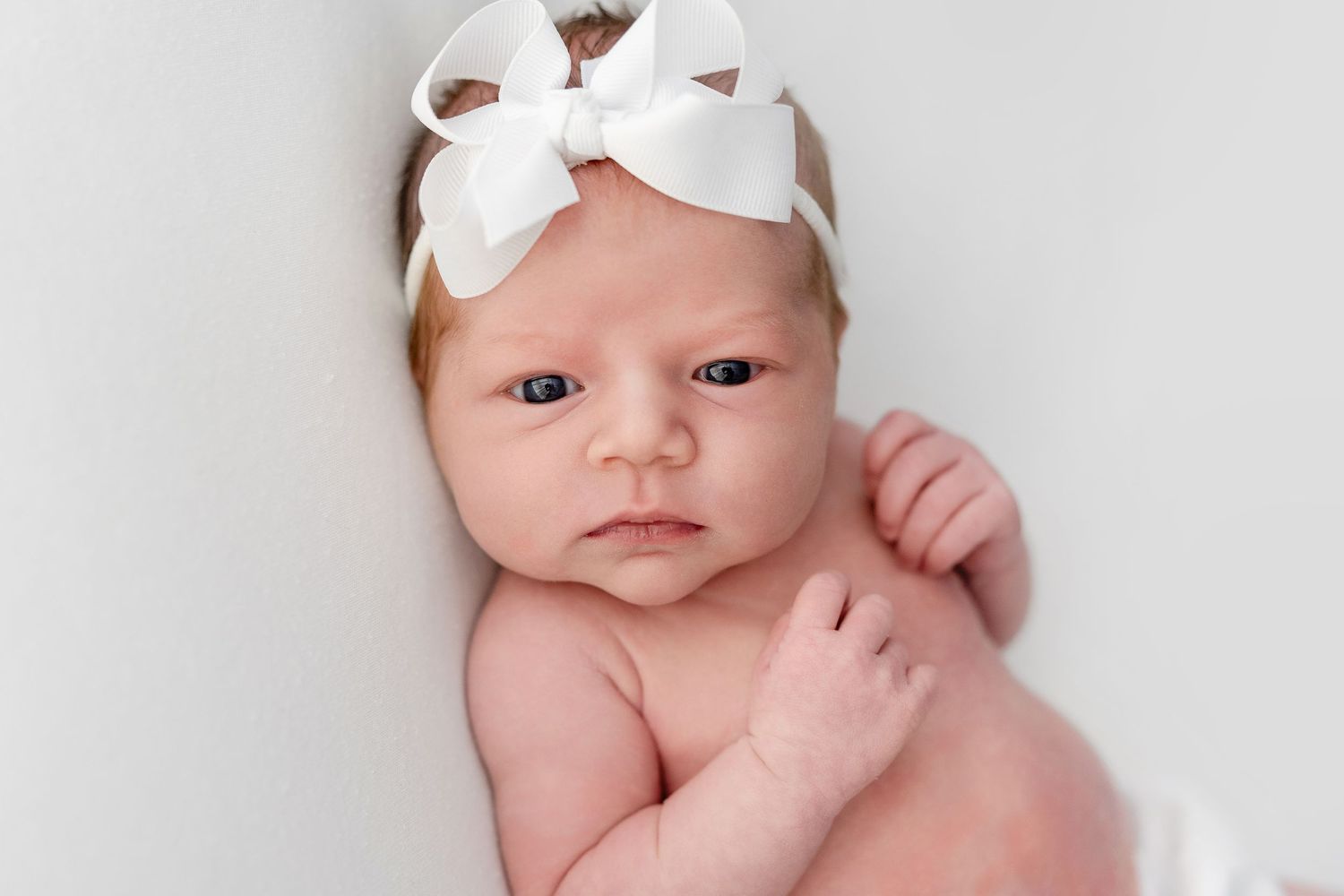 Close-up portrait of a newborn baby wearing a white bow headband against a light background.