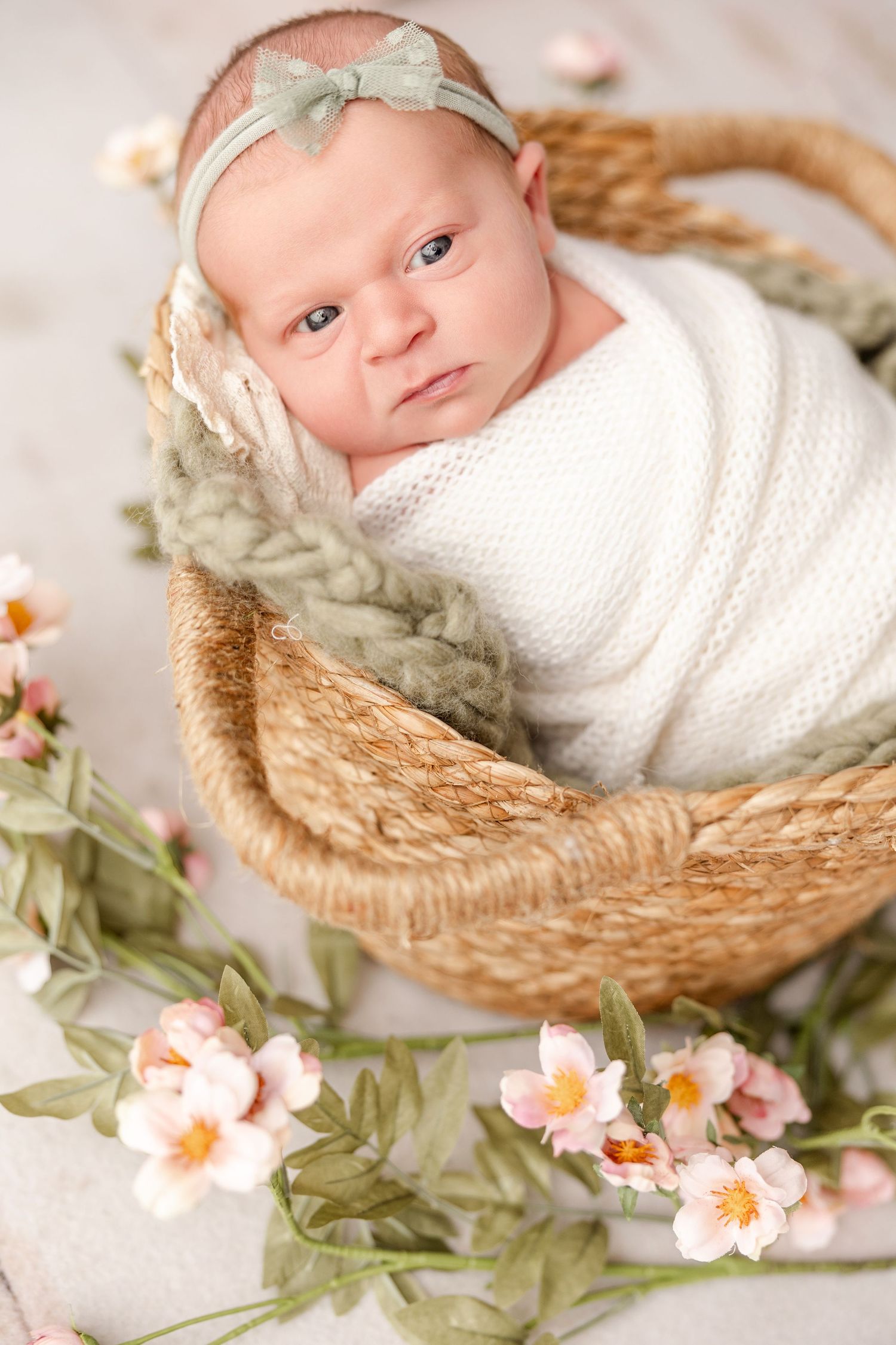 Newborn baby wearing white swaddle rests peacefully in woven basket surrounded by delicate pink flowers and greenery.