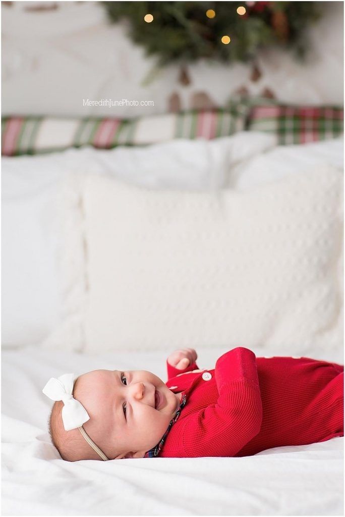 Baby in red holiday outfit lying on white bedding with Christmas tree and lights in background.