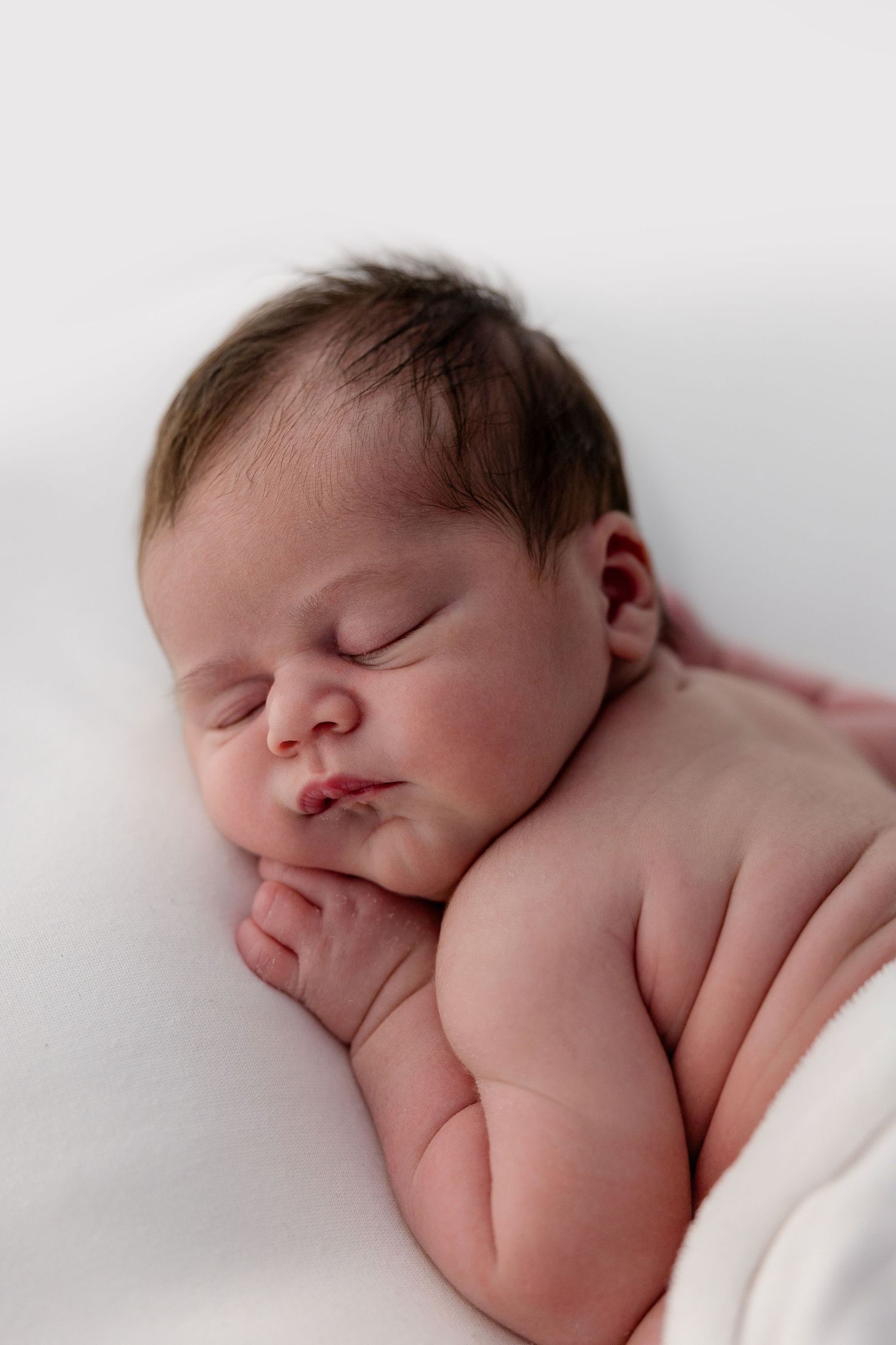Peaceful newborn baby sleeping on a white blanket with dark hair and tiny hands tucked under chin.