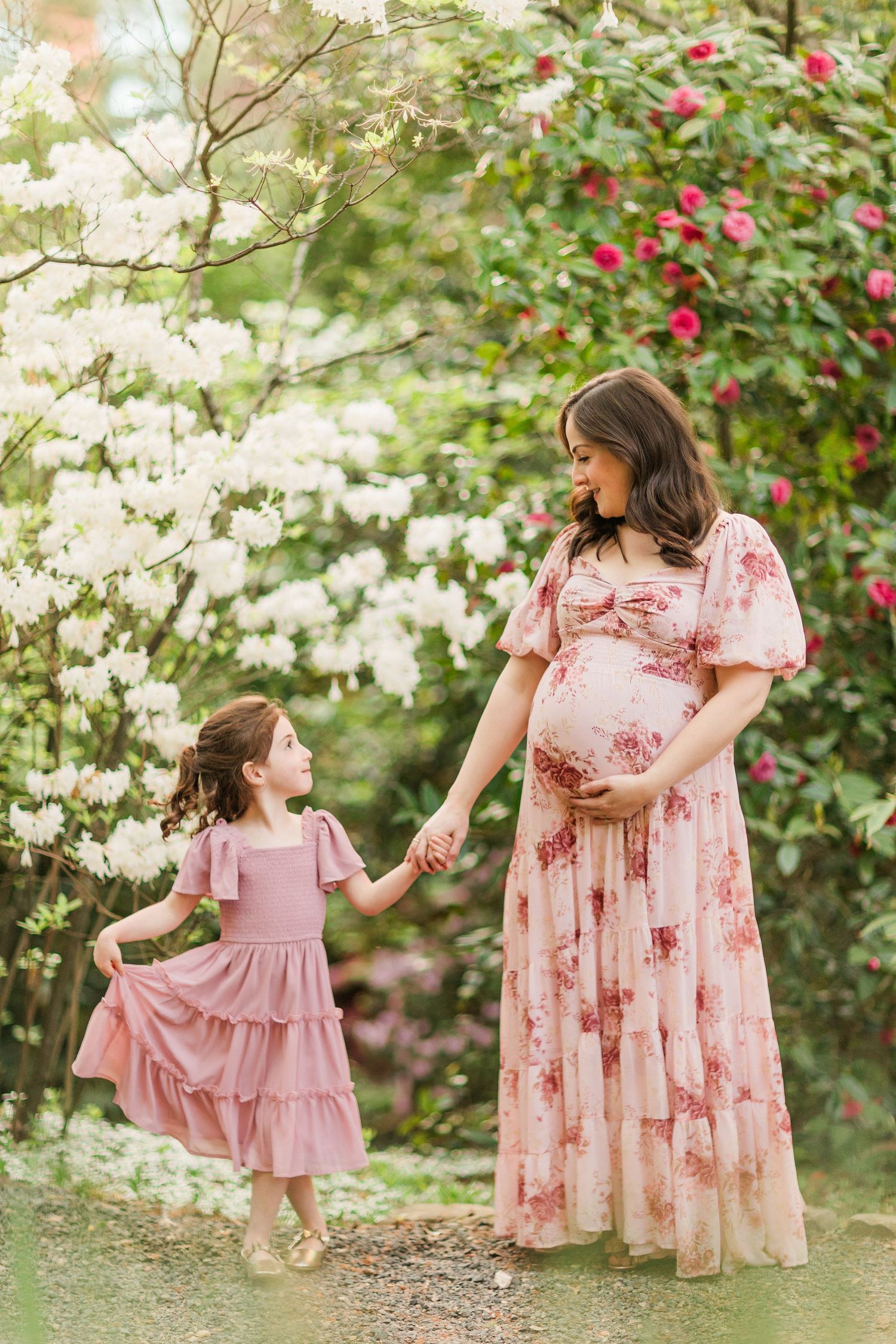 Mother and daughter hold hands in matching pink dresses among blooming spring flowers in a garden setting.