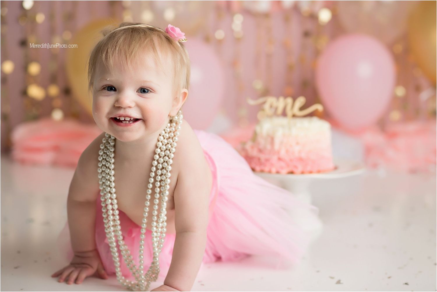Baby in pink tutu and pearls poses at first birthday photoshoot with cake and balloons in soft pink setting.