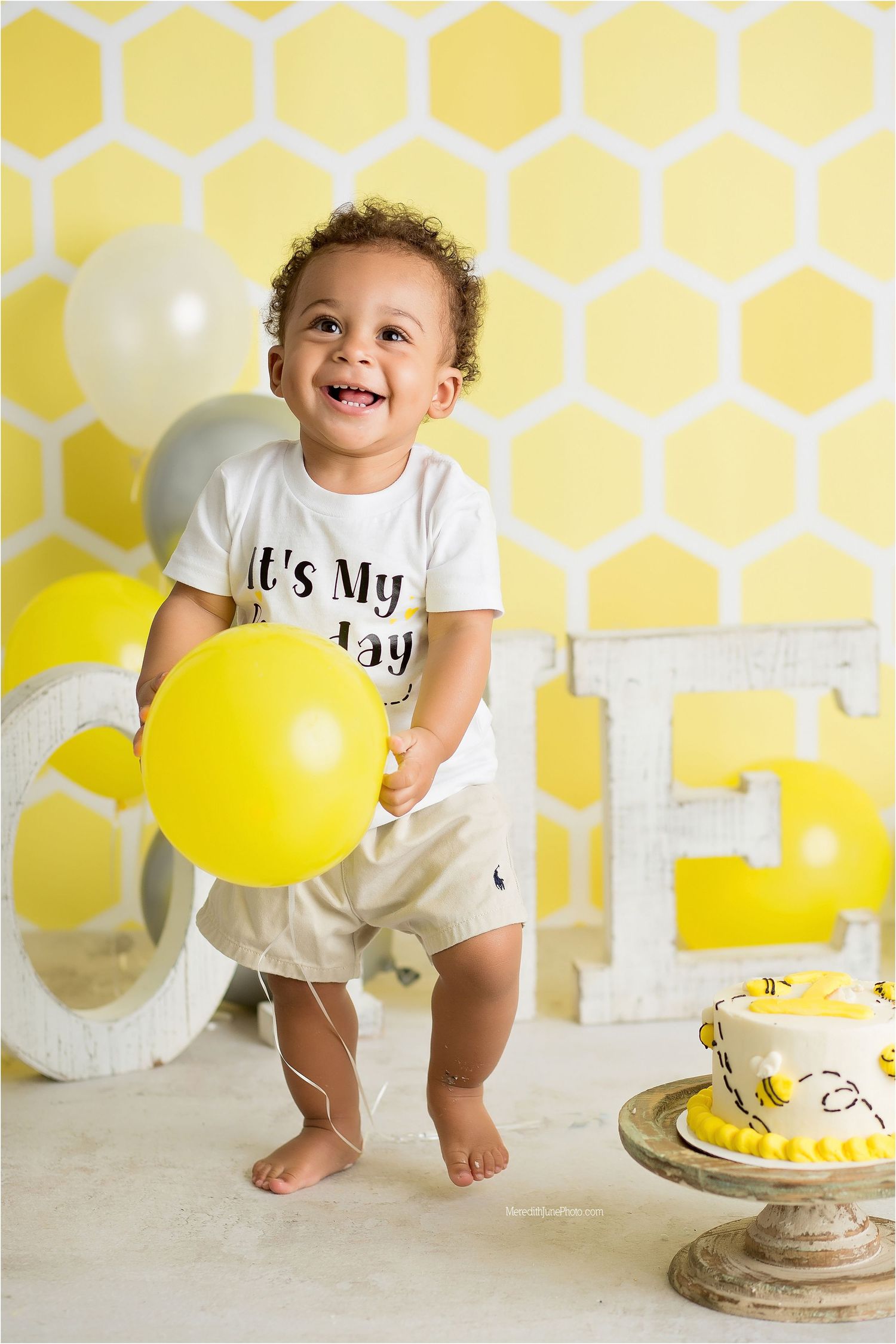 A joyful celebration scene with yellow balloons, honeycomb wall pattern, and a birthday cake on a pedestal.