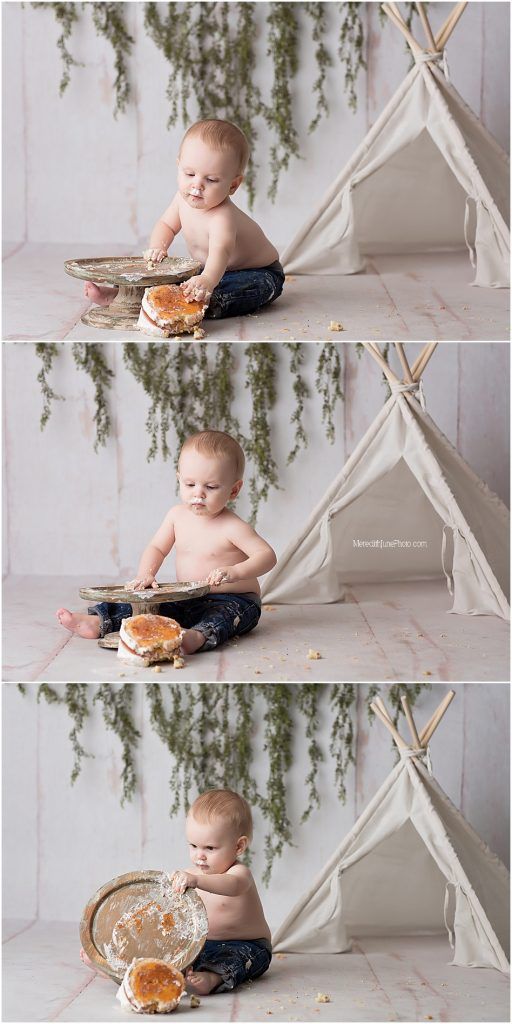 Baby in dark pants explores a cake smash photo session with white teepee and greenery backdrop in studio setting.