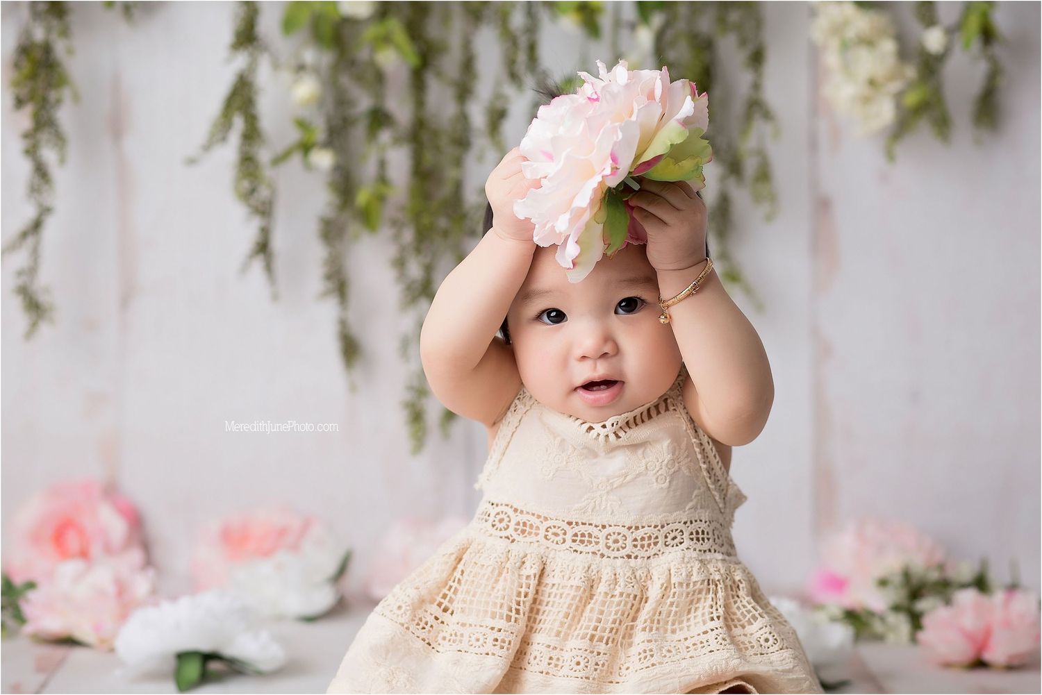 Baby wearing a cream crochet dress sits in front of a floral backdrop during a spring photo session.