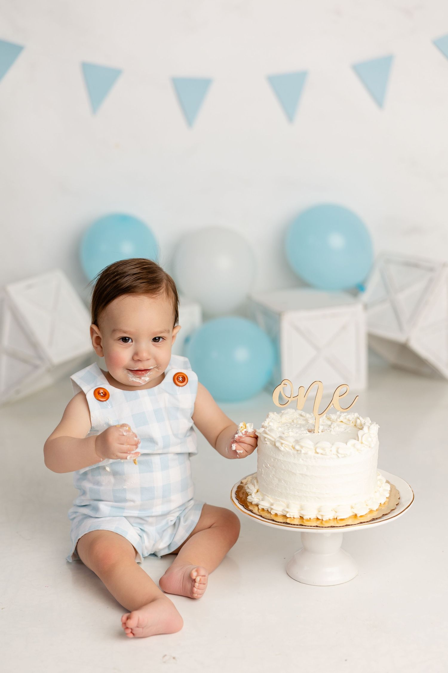 Baby in light blue outfit sits near white birthday cake with blue balloon and bunting party decorations.