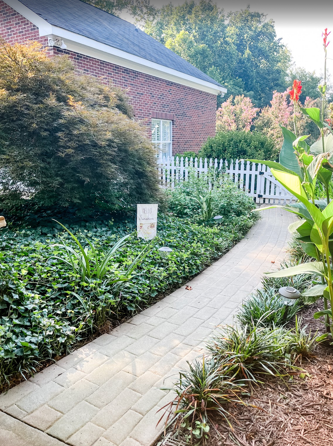 A curved concrete pathway winds through a landscaped garden with brick buildings and white picket fencing in the background.