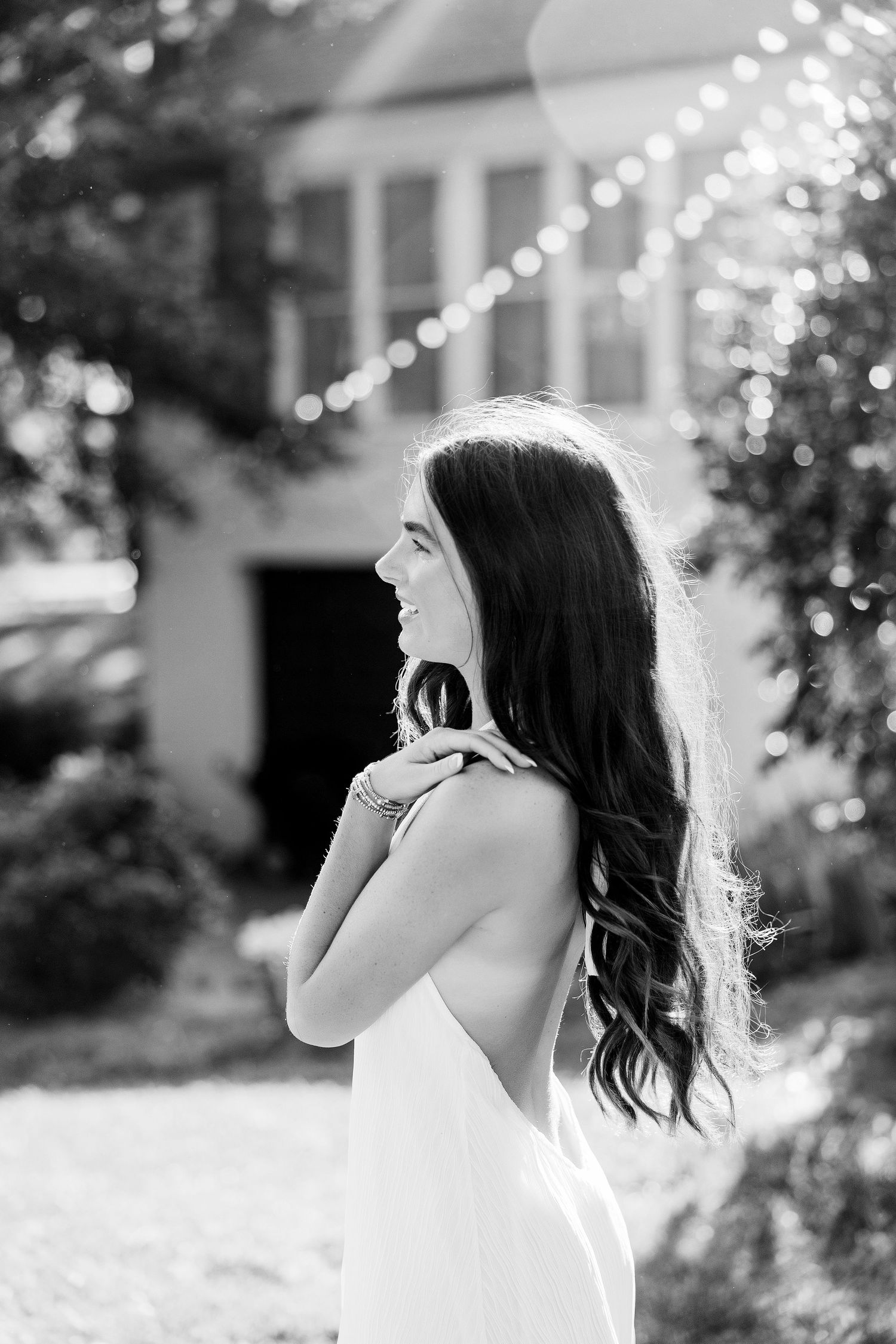 Black and white photo of a smiling bride in a backless gown, standing outdoors with bokeh lights and greenery.