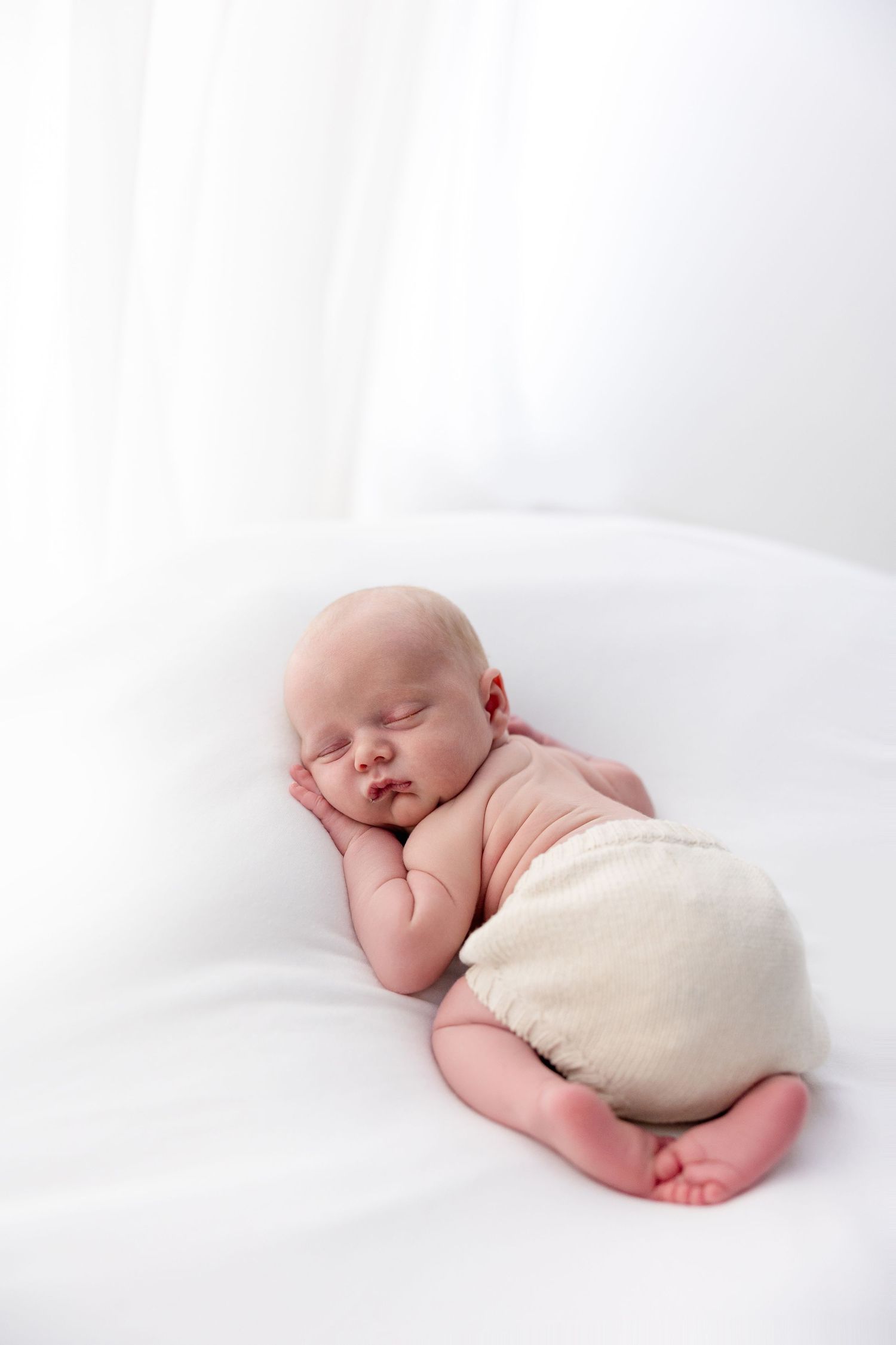 Peaceful newborn baby sleeping on white blanket wearing cream diaper cover in professional studio portrait.