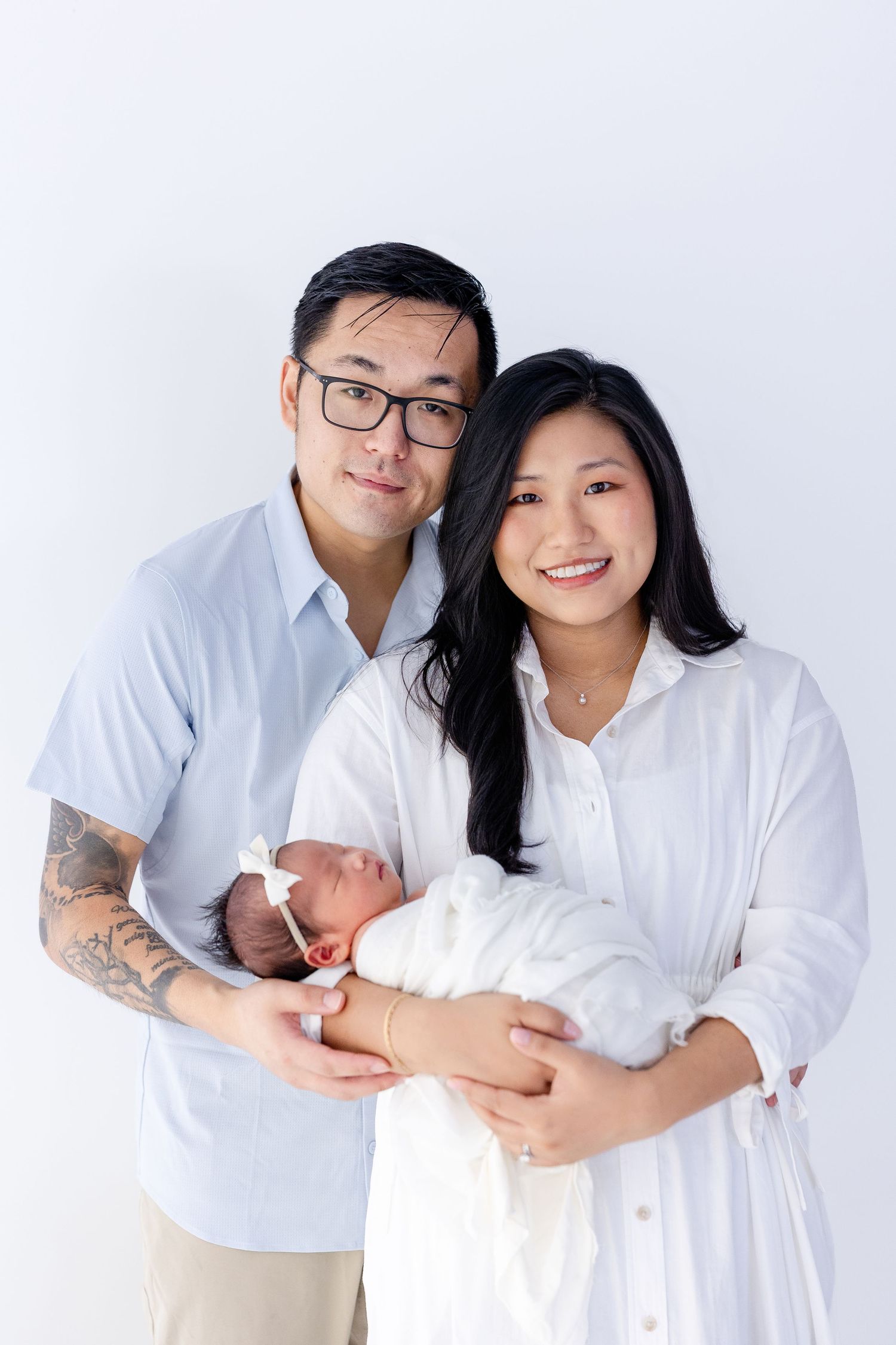 Happy Asian couple smiling and holding their swaddled newborn baby against a white background.