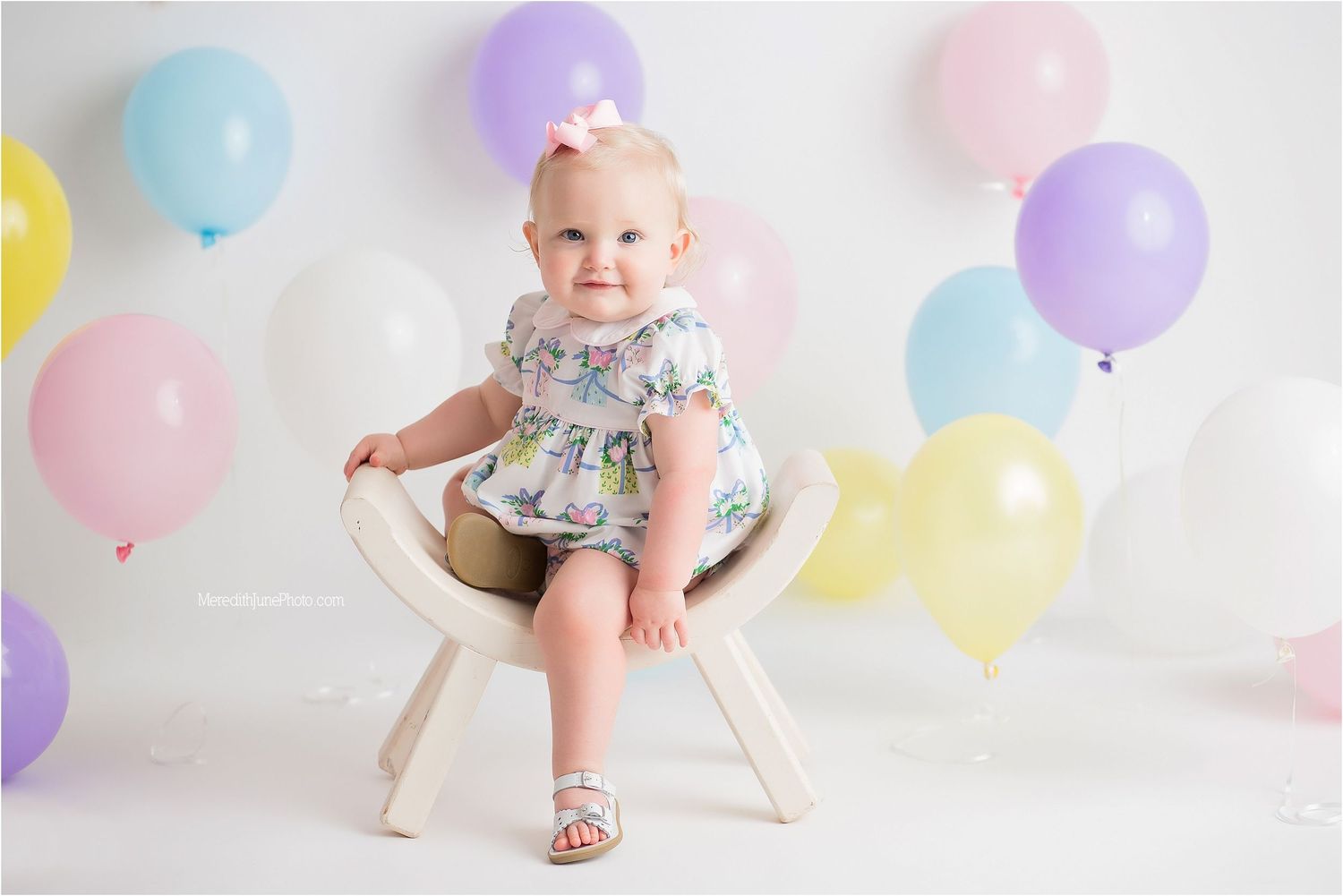 Small child sits on white chair surrounded by pastel balloons in pink, yellow, purple and blue against white backdrop.