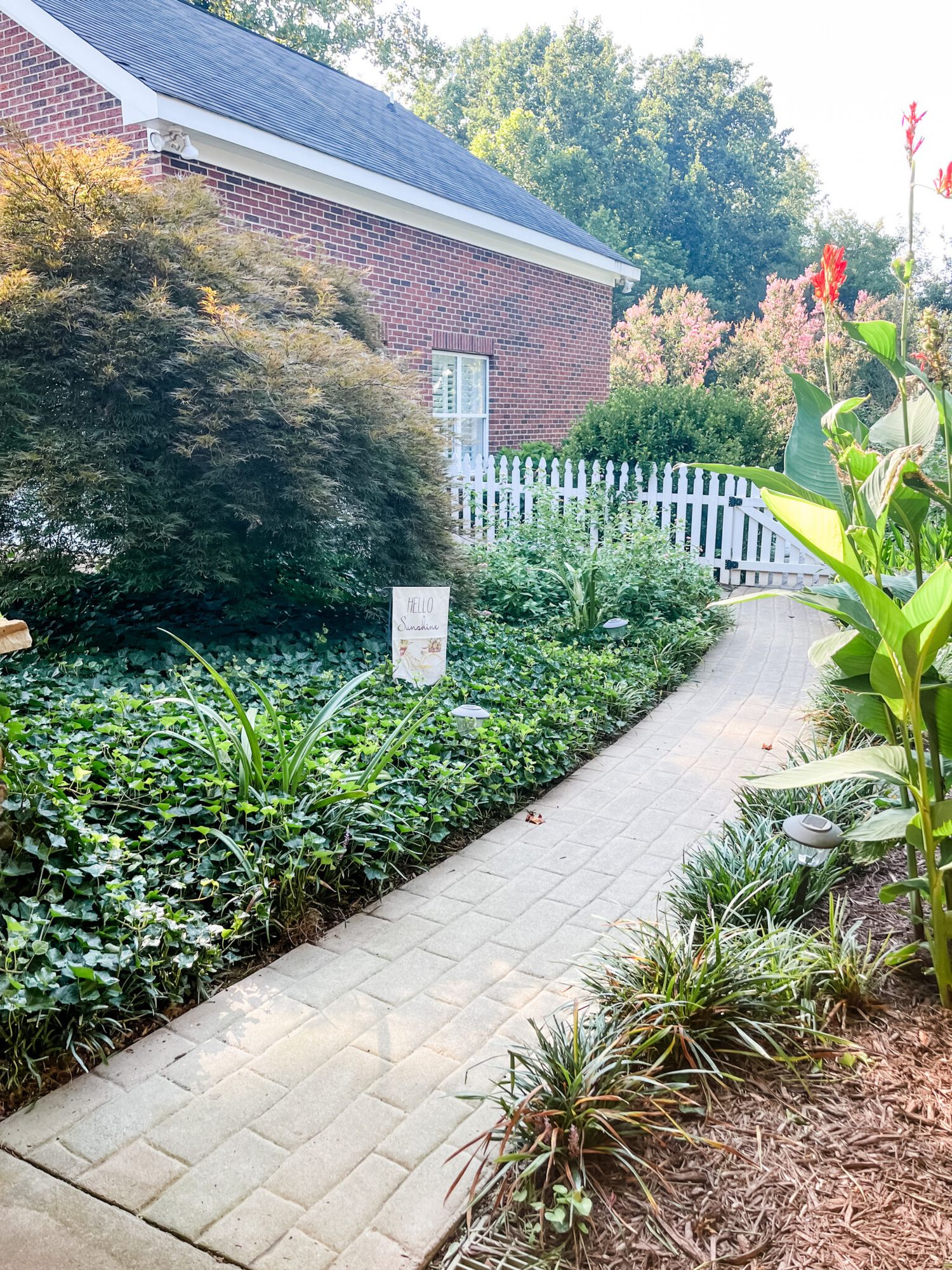 A curved stone pathway leads through a landscaped garden with shrubs and tropical plants beside a brick building.