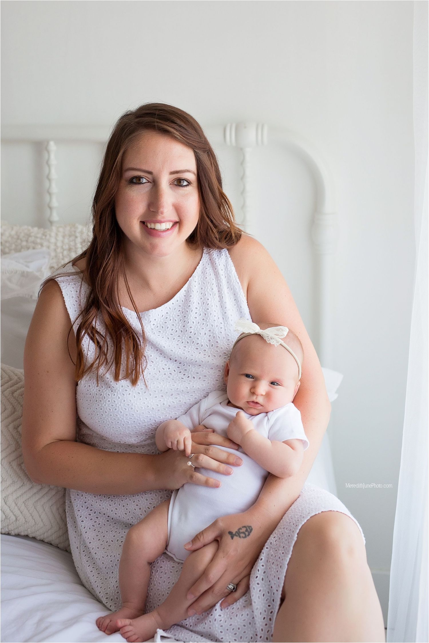 A baby in white clothing sits on a bed with decorative pillows in a bright room.