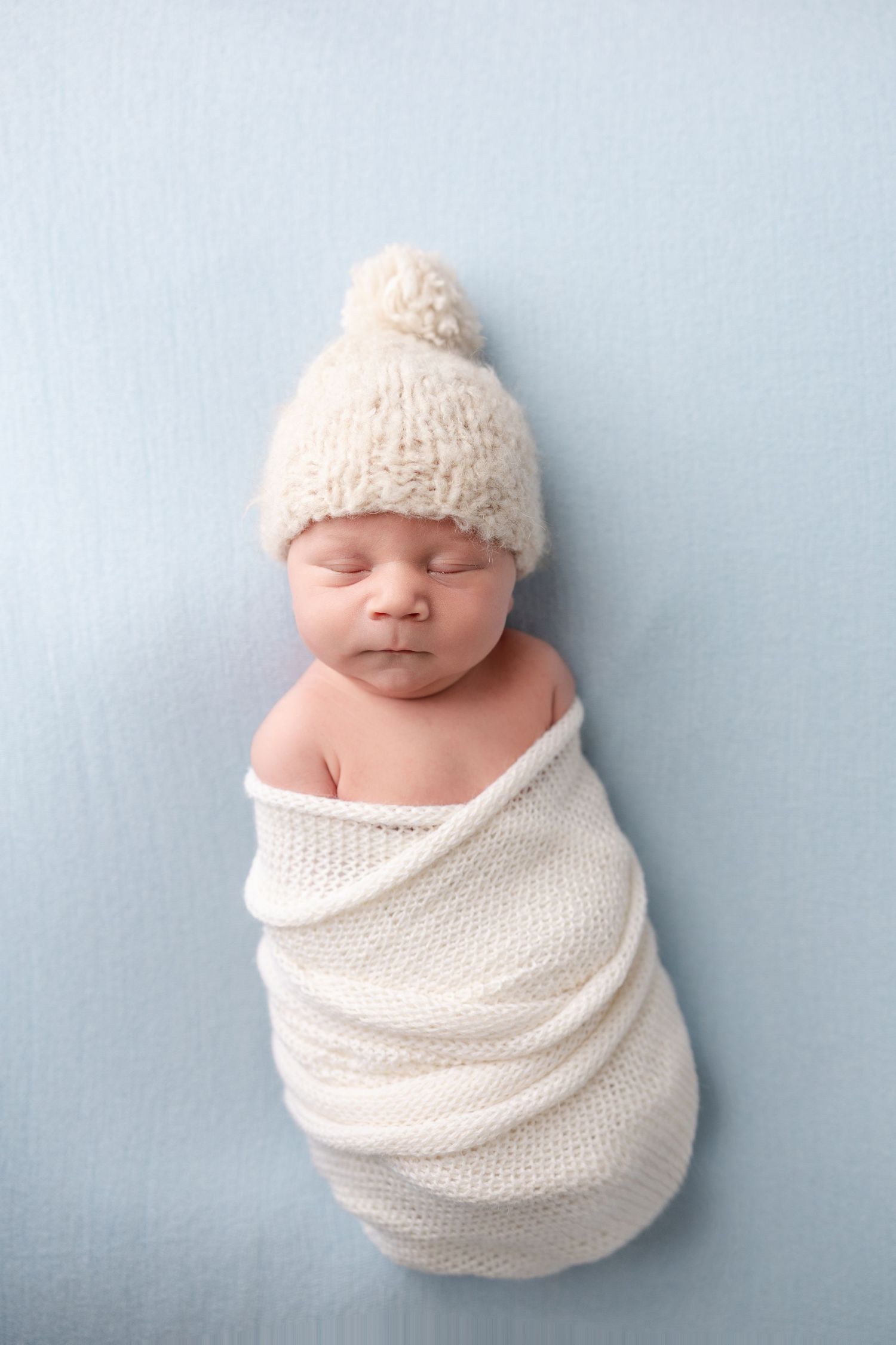A sleeping newborn baby wrapped in soft white knit fabric against a light blue background.