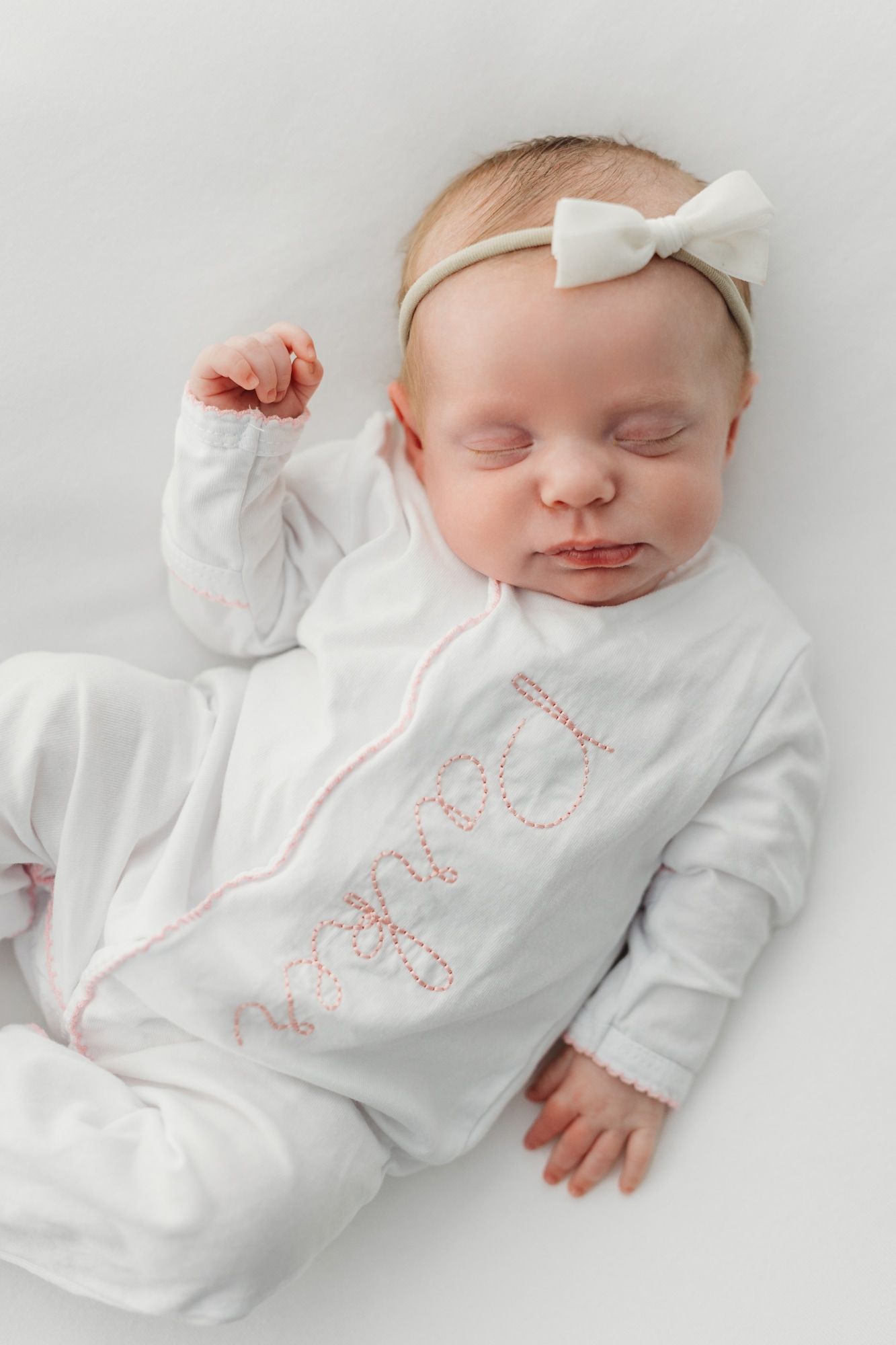 Sleeping newborn in white outfit with bow headband on light background.