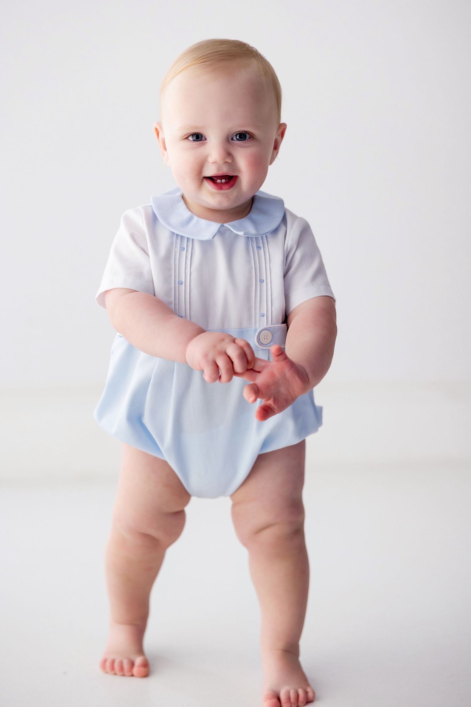 Baby wearing light blue and white formal outfit sitting on white backdrop.