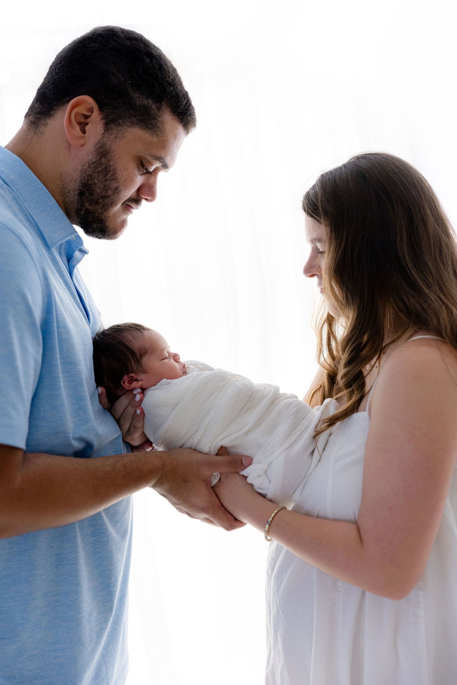 New parents lovingly gaze at their swaddled newborn baby in a bright, airy studio portrait.