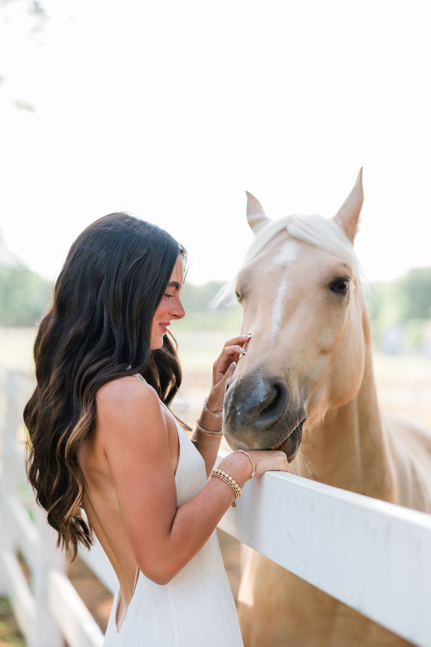 Young woman with long dark hair in a white dress gently petting a palomino horse at a white fence.