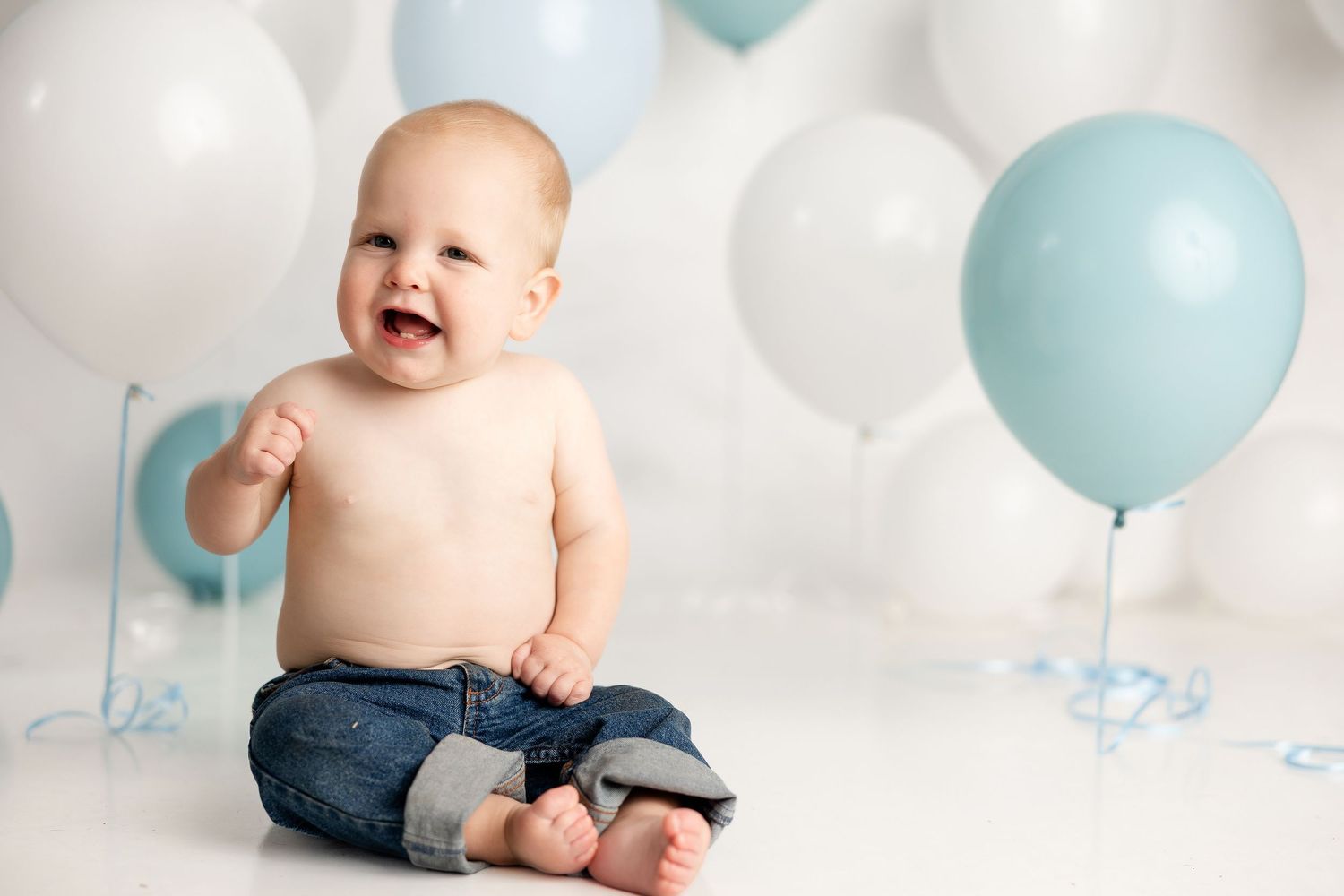 Baby sits among white and blue balloons in casual denim outfit during milestone photoshoot.