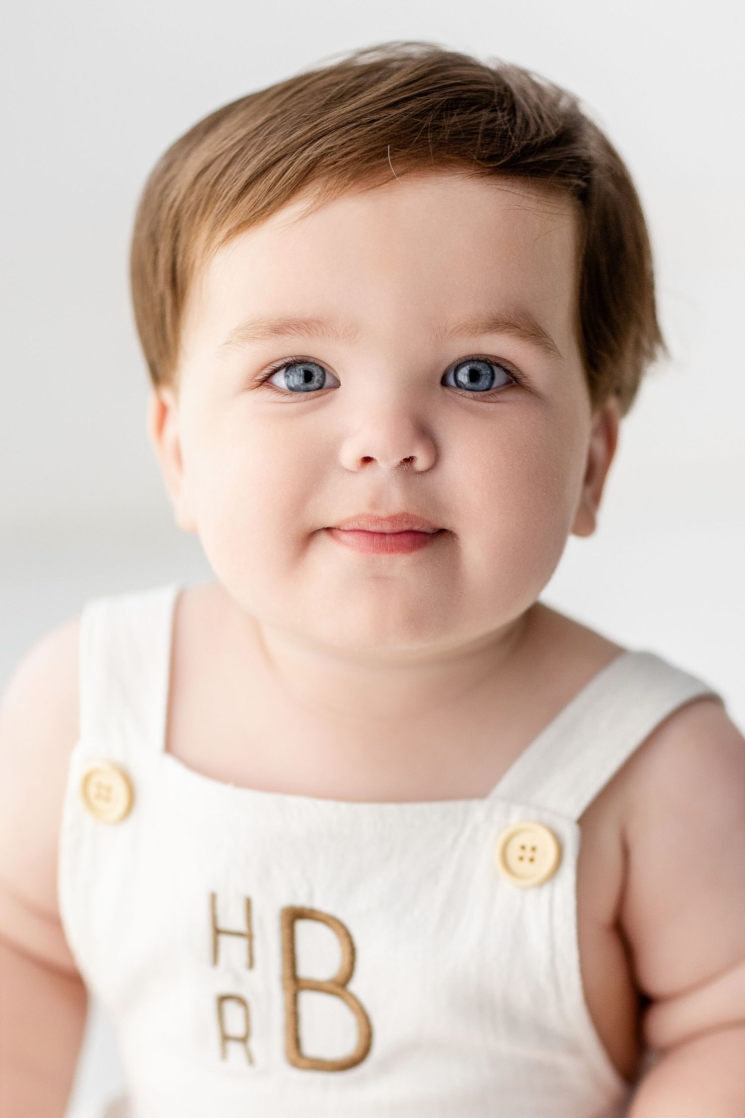 Baby wearing white outfit with buttons smiles at camera in studio portrait.