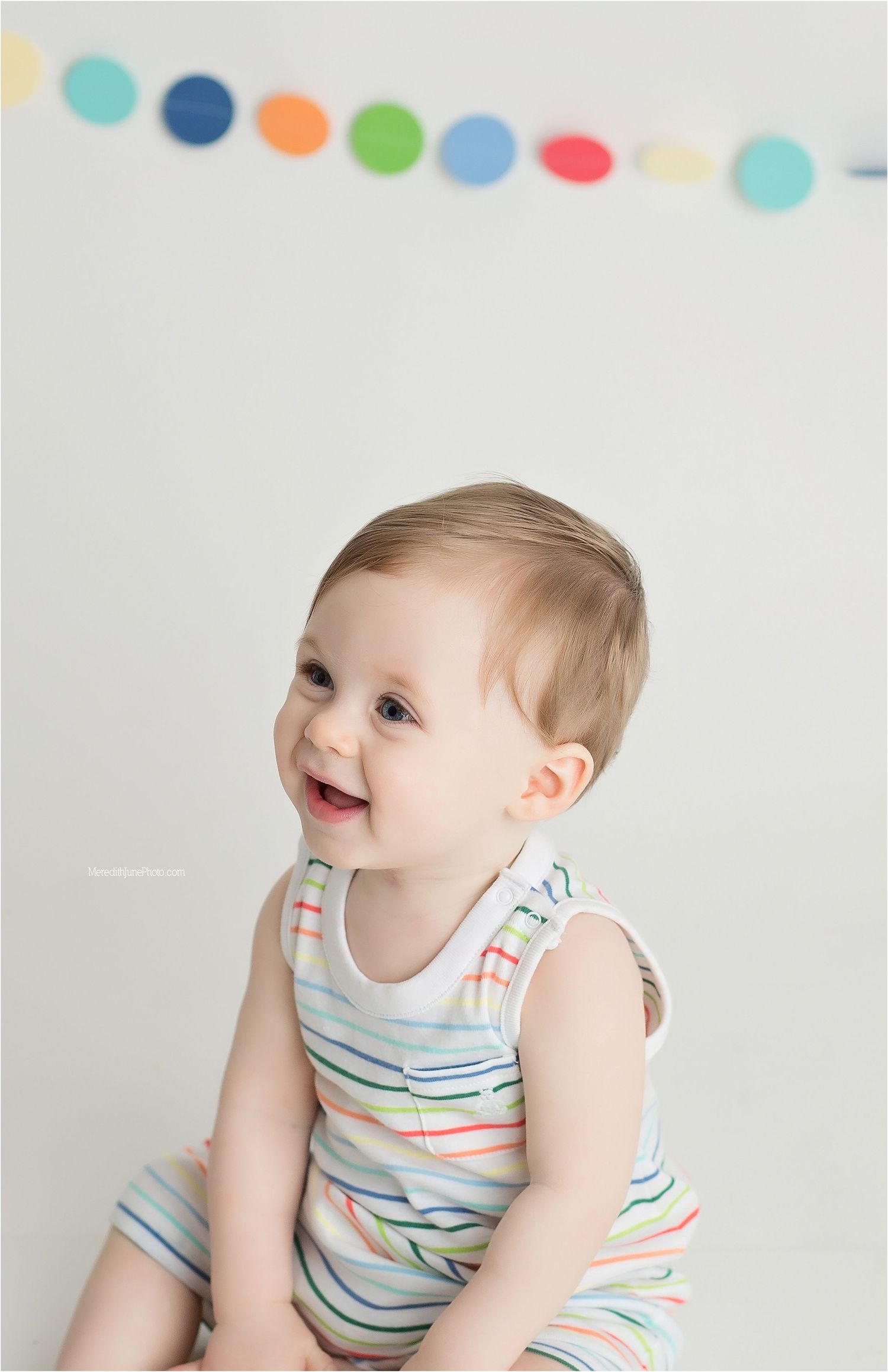 Happy baby wearing striped romper sitting against white wall with colorful polka dot banner decoration.