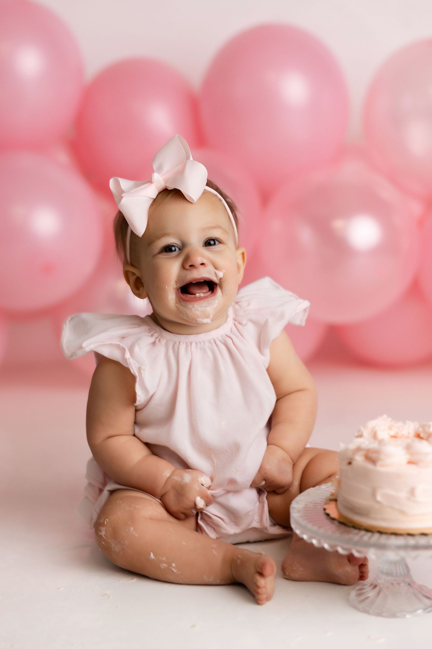 Baby in pink dress sits on white backdrop with pink balloons and cake during first birthday photo session.