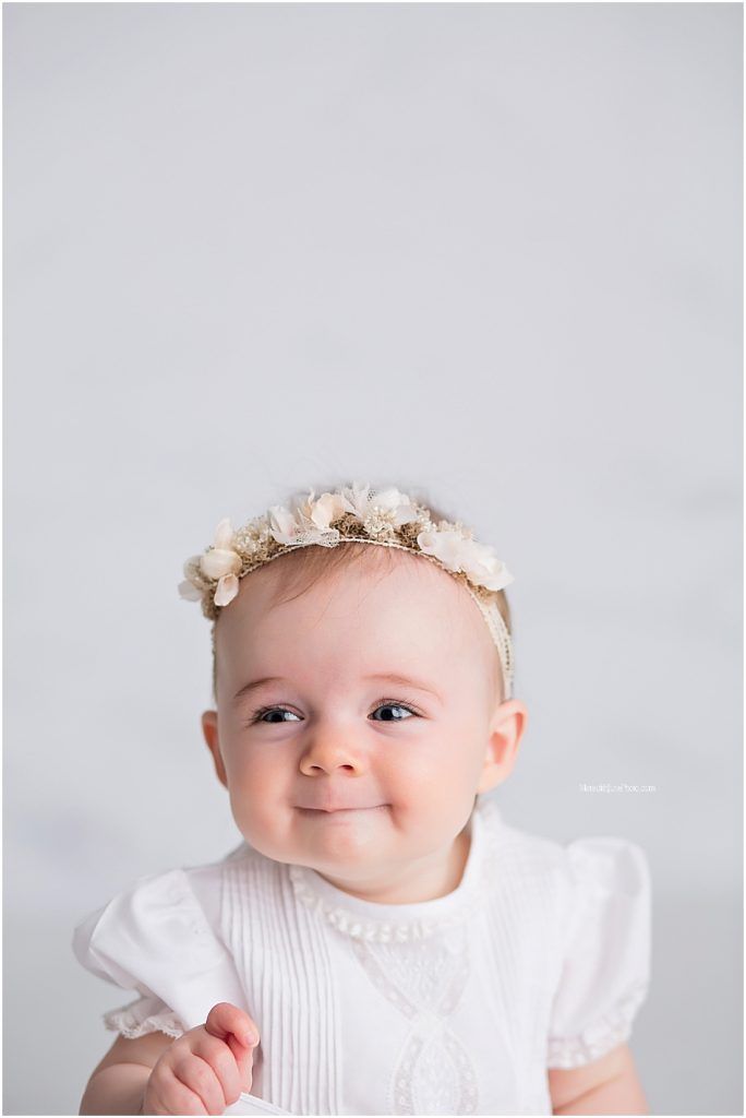 A baby wearing a white dress and delicate floral crown poses against a light gray background.