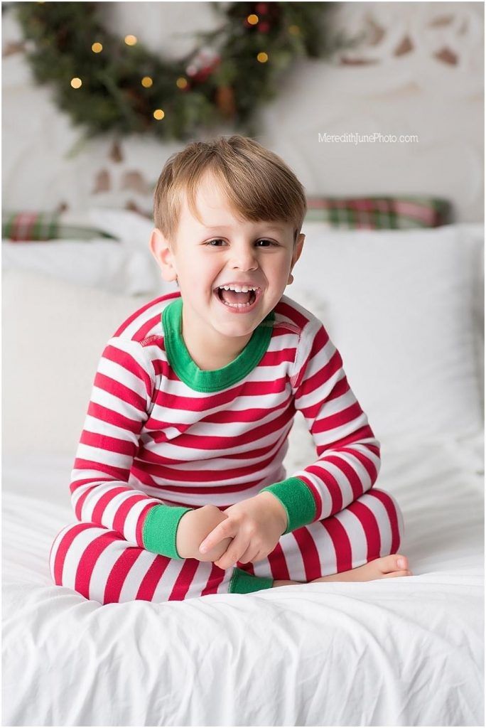 Child wearing red and white striped holiday pajamas lying on white bedding with Christmas decorations in background.
