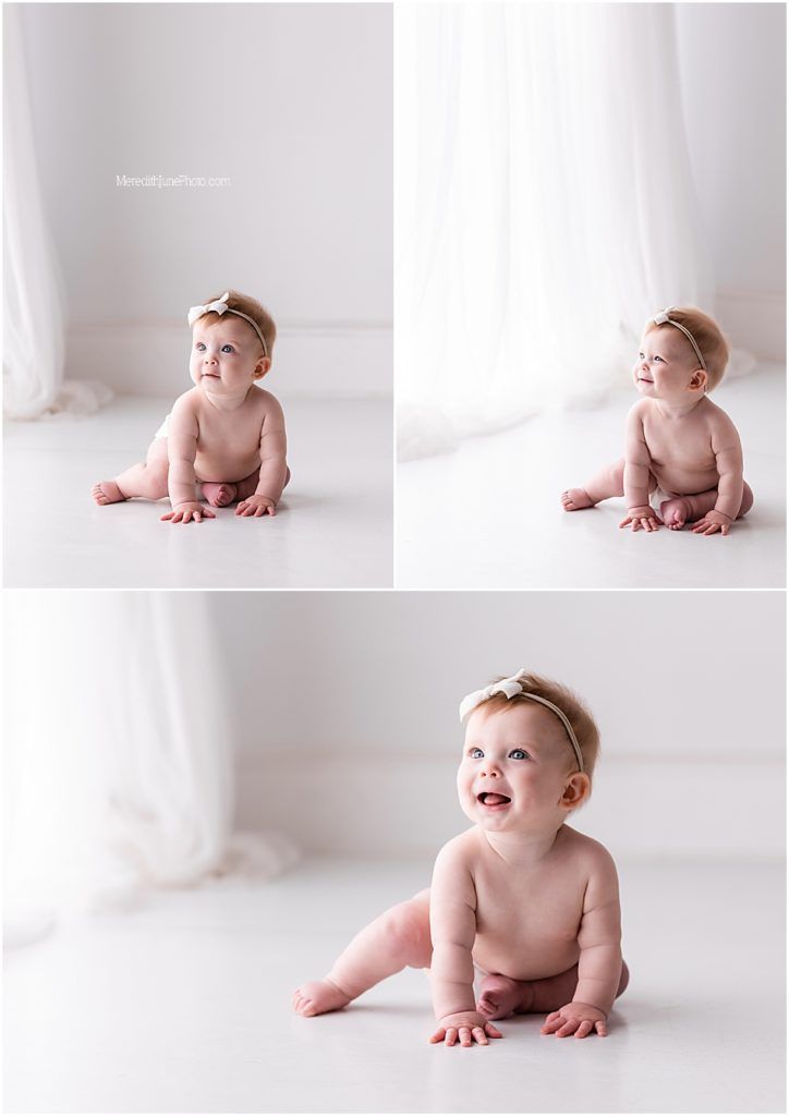 A baby in white surroundings sits and smiles while wearing a small bow headband in a bright minimalist studio setting.