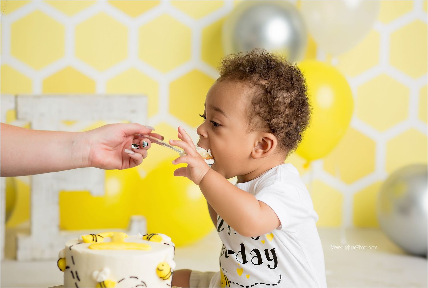Baby in a high chair enjoying a first birthday cake smash photoshoot against a yellow honeycomb backdrop.